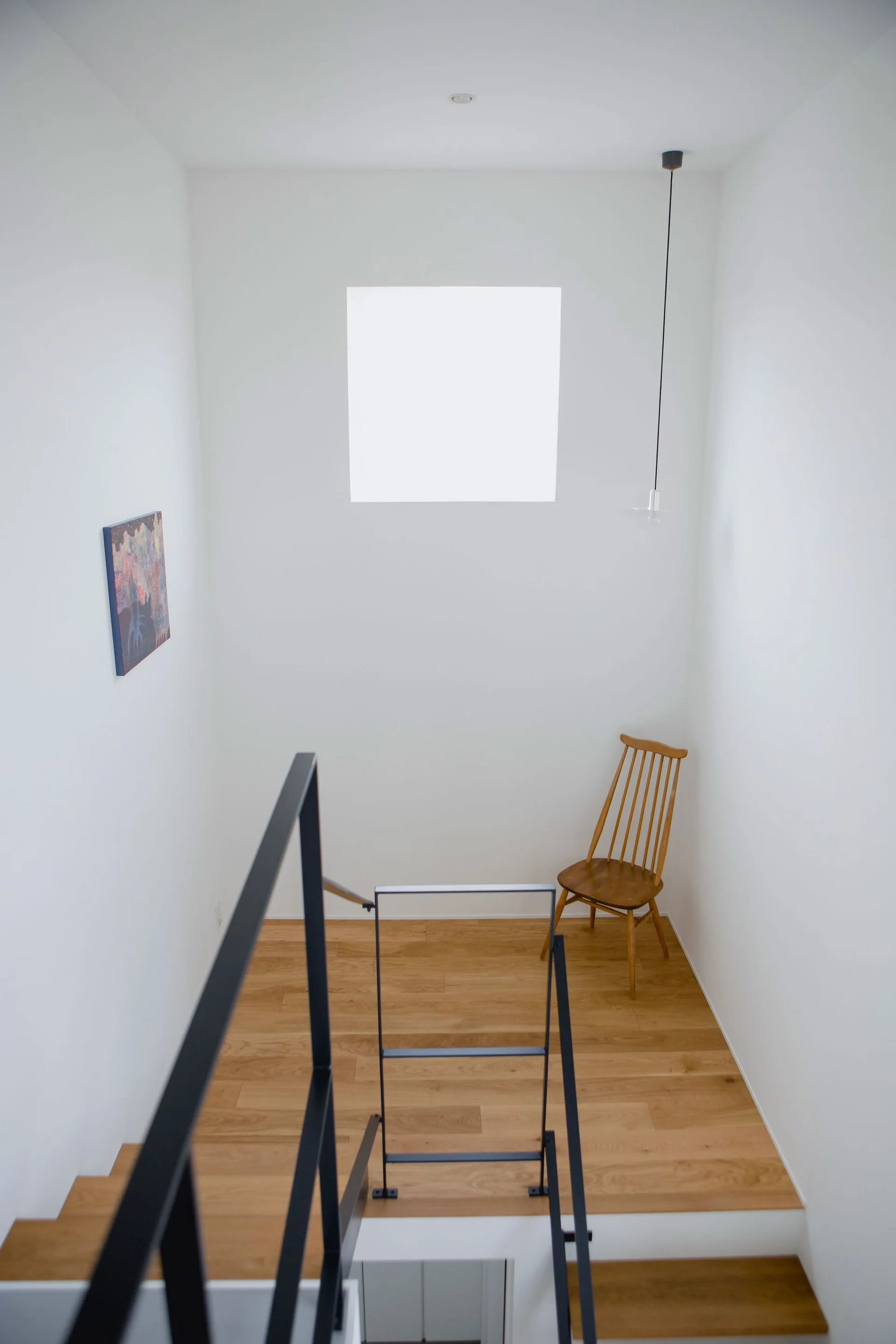 Looking down a white stairwell with wood steps, black metal railings, a square window, and a single wooden spindle-back chair on the landing.