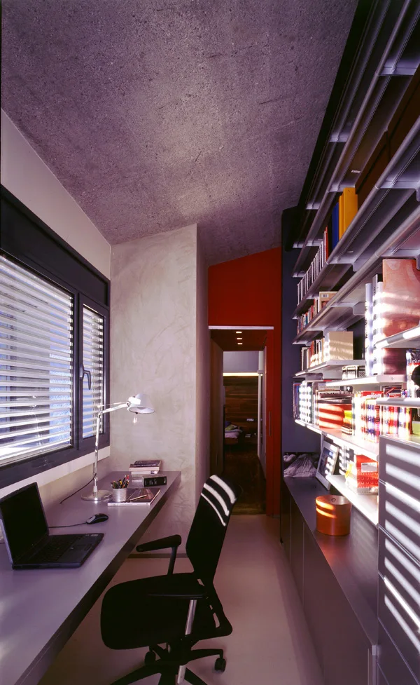 A narrow home office features a long wall-mounted grey desk, a black office chair, metal track shelving, and a raw concrete ceiling.