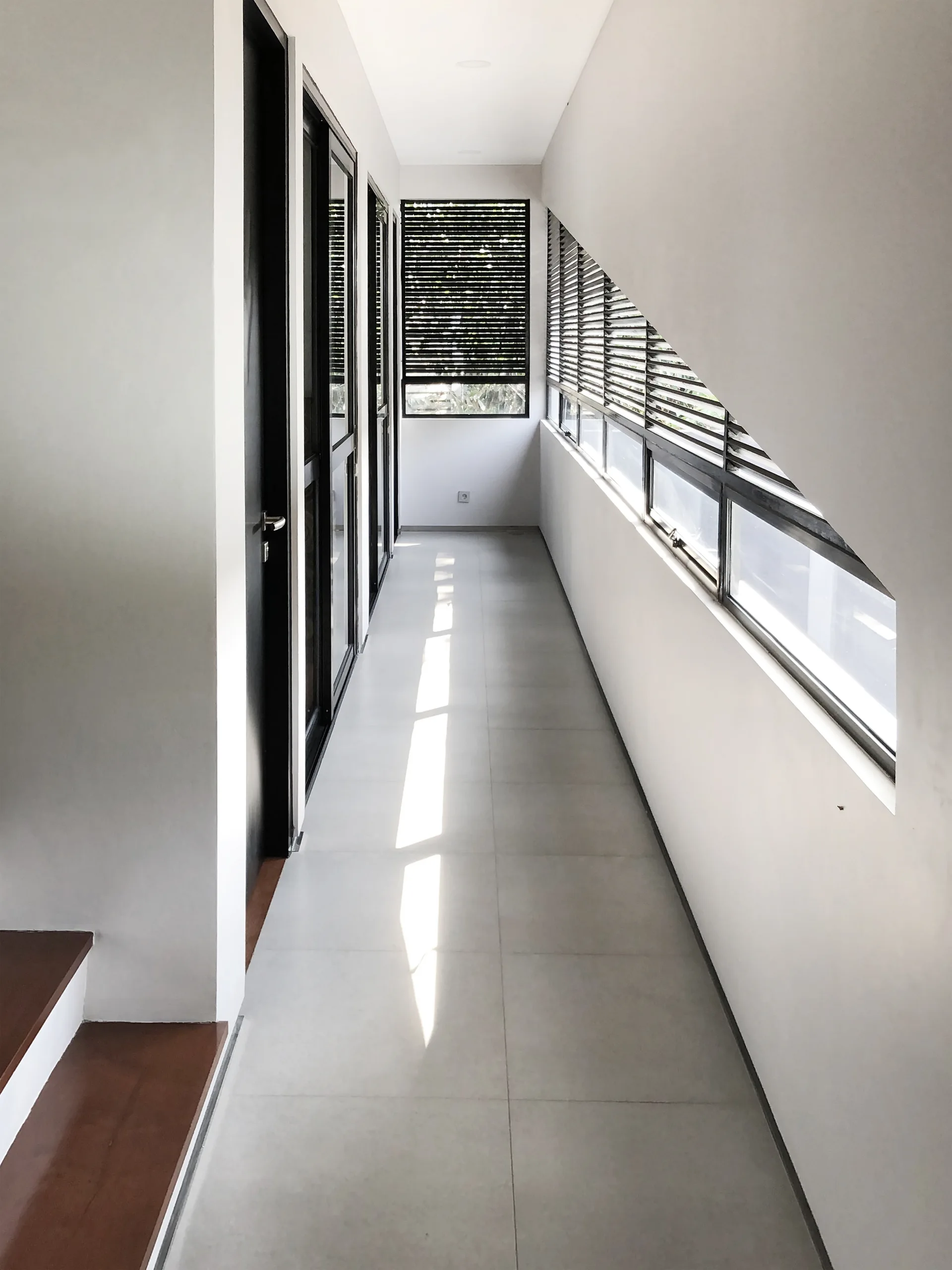 A minimalist white hallway with light grey floor tiles, black-framed glass doors, and a slanted window wall with horizontal blinds.