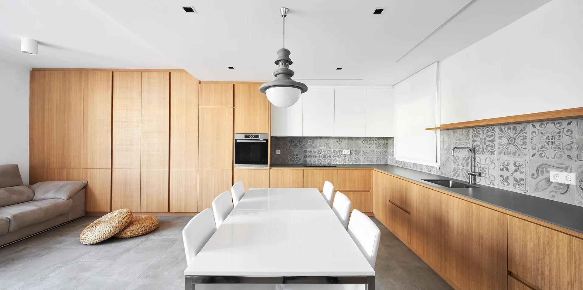 A modern open-concept kitchen features wood cabinetry, a white rectangular dining table, a grey patterned tile backsplash, and concrete floors.