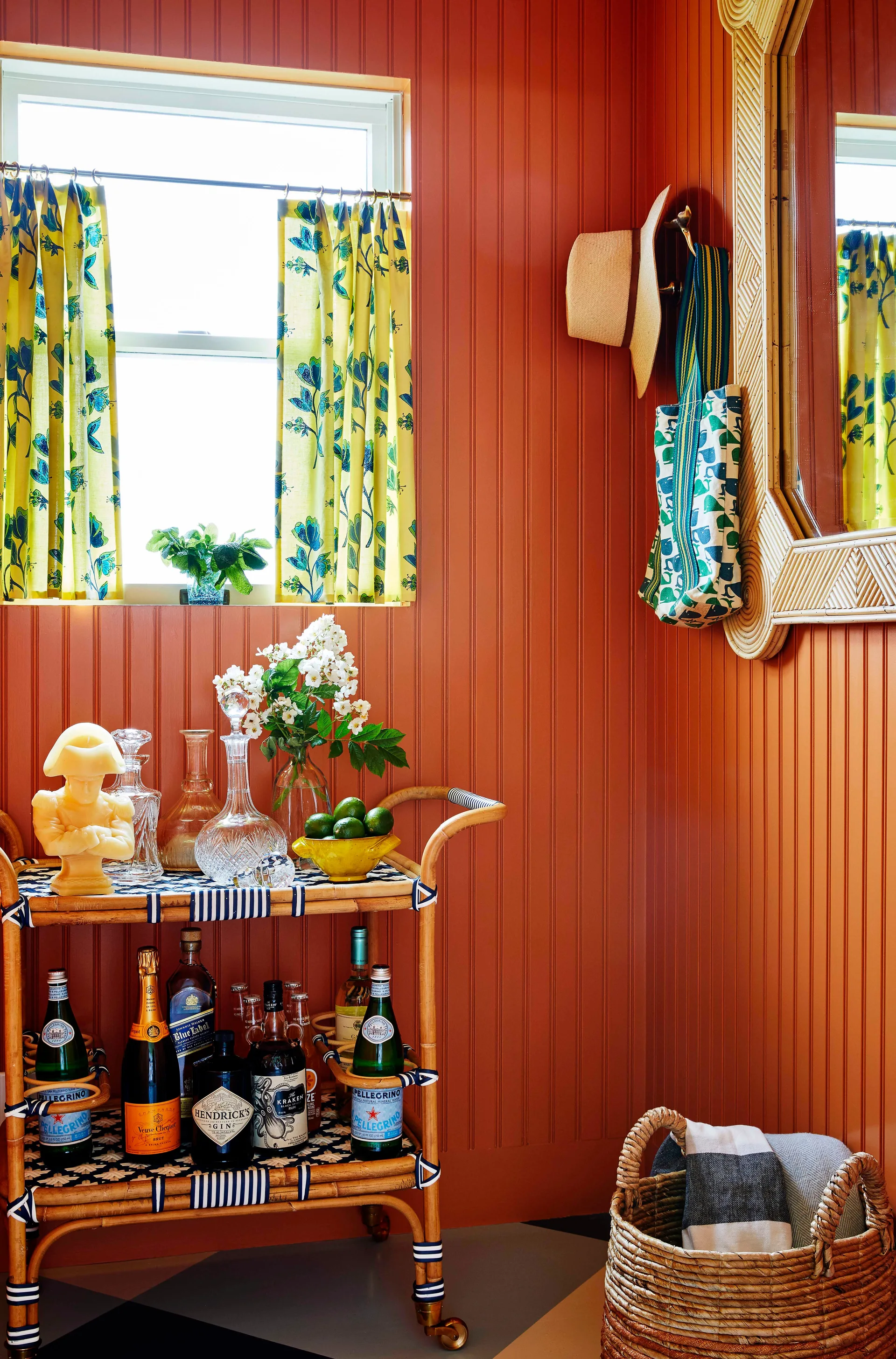 A terracotta beadboard room features a rattan bar cart stocked with bottles, a window with yellow floral cafe curtains, and a rattan mirror.