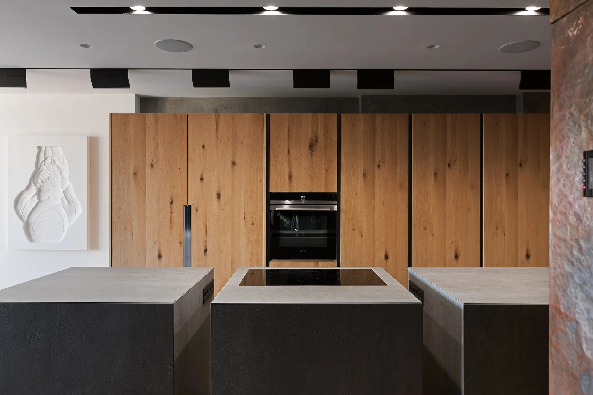 A minimalist kitchen with a tall wall of wood flat-front cabinets and three monolithic grey island blocks under recessed track lighting.
