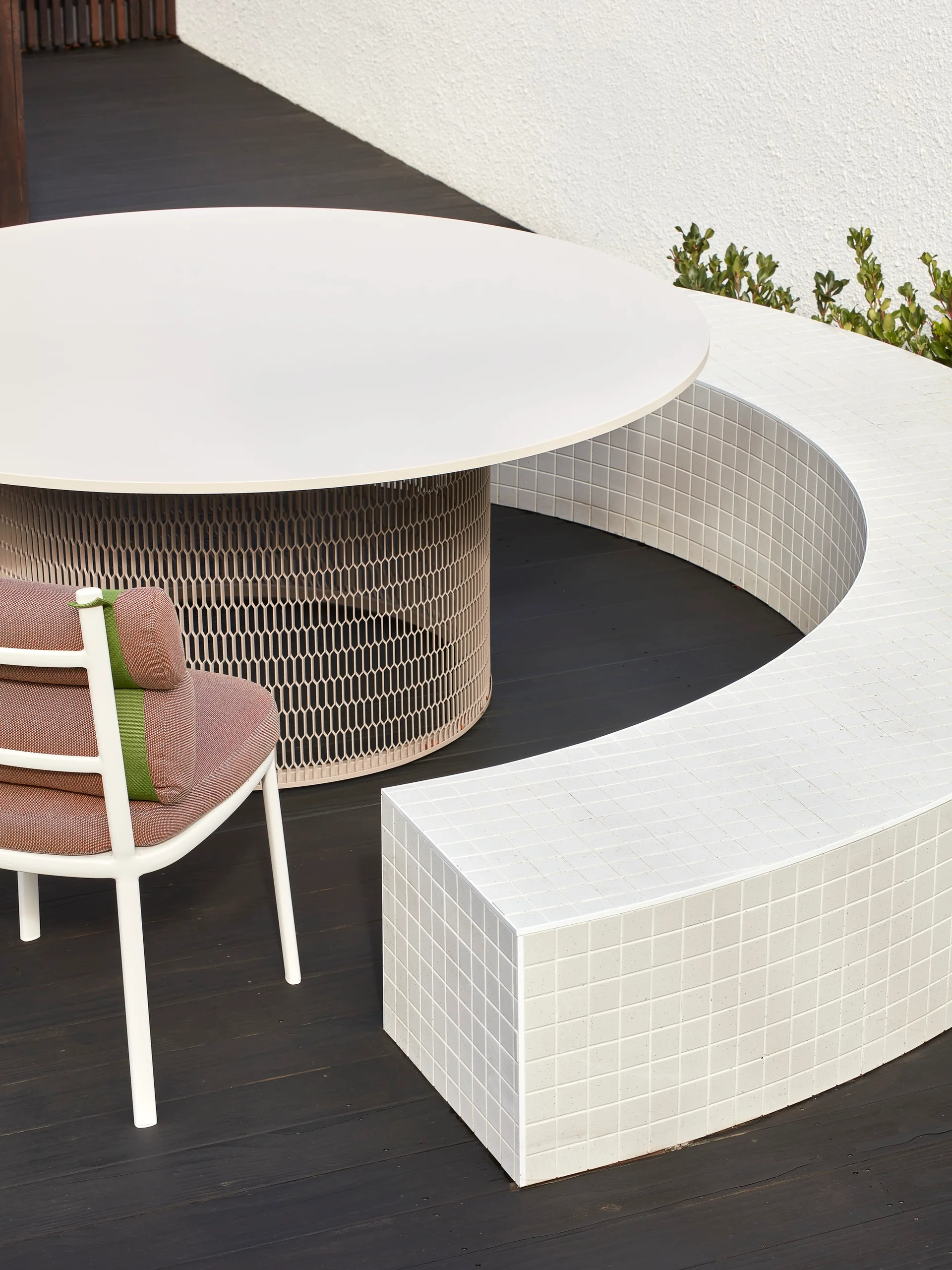 A modern outdoor patio features a round white dining table and a curved built-in bench covered in small white tiles on a dark wood deck.