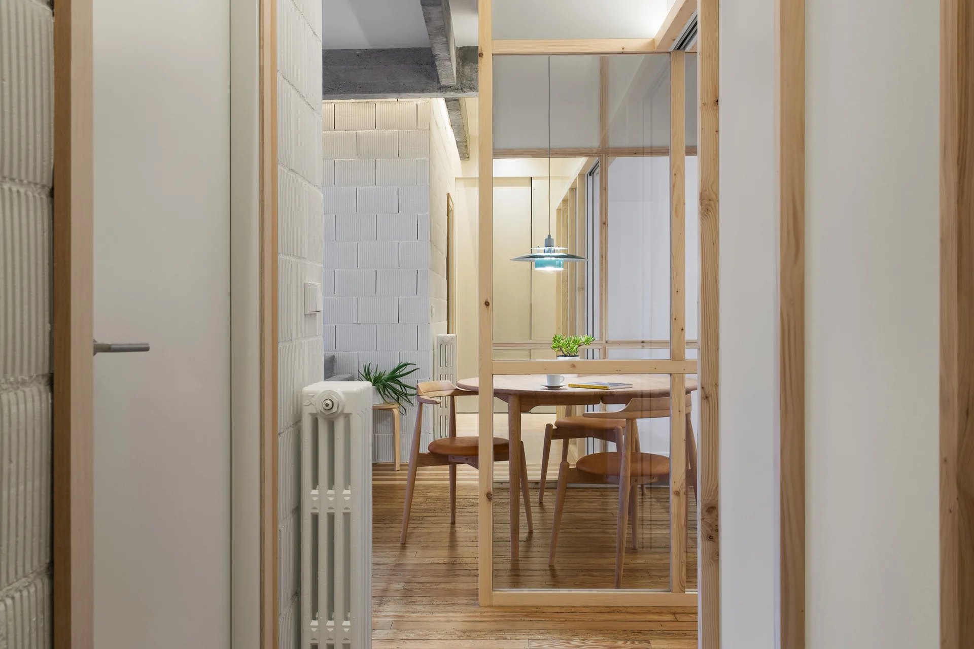A view through a light wood and glass partition into a dining room with a wooden table, mid-century chairs, and white block walls.
