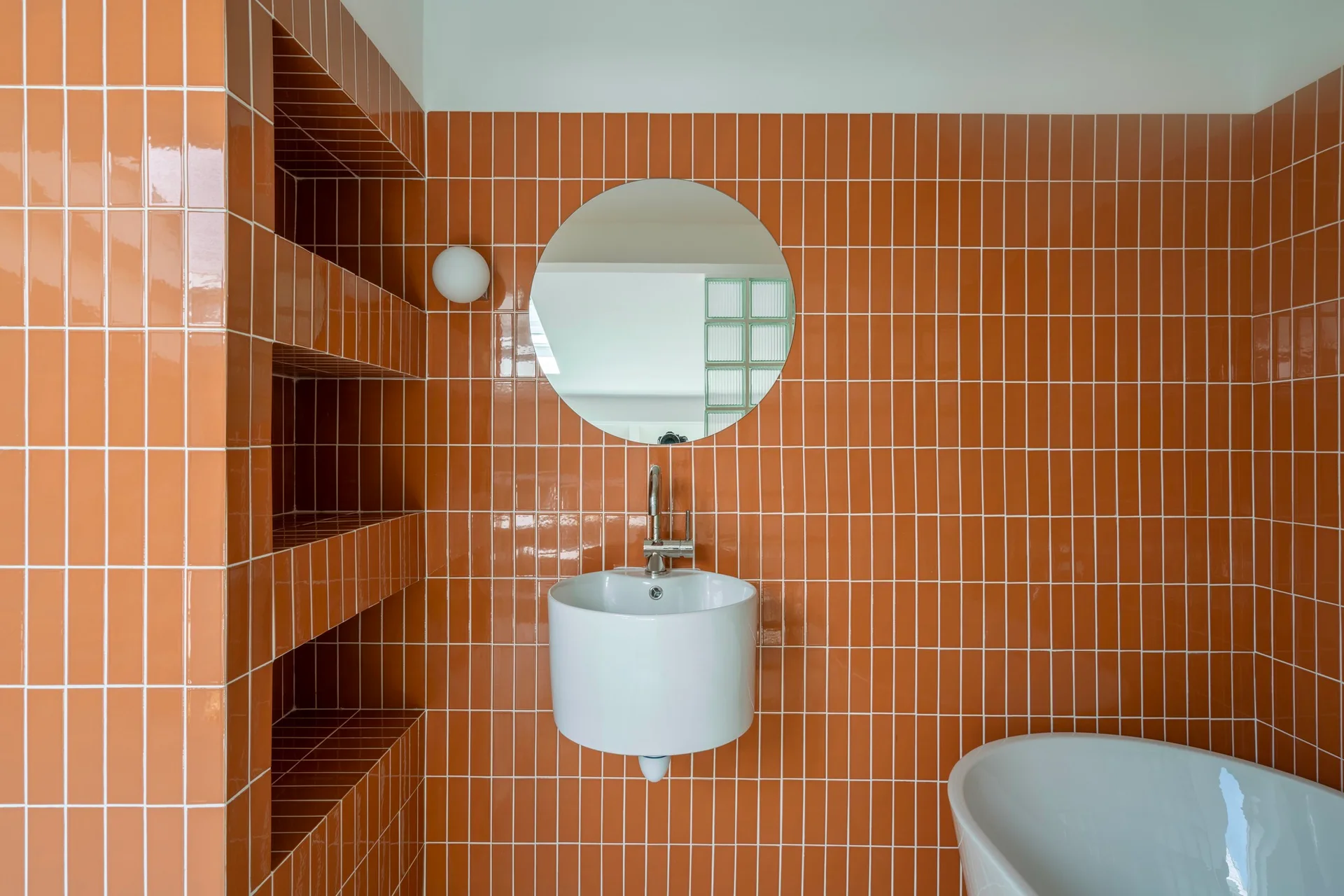 A minimalist bathroom features bright orange vertical tiles on the walls, a circular mirror, a white wall-mounted sink, and recessed shelving.