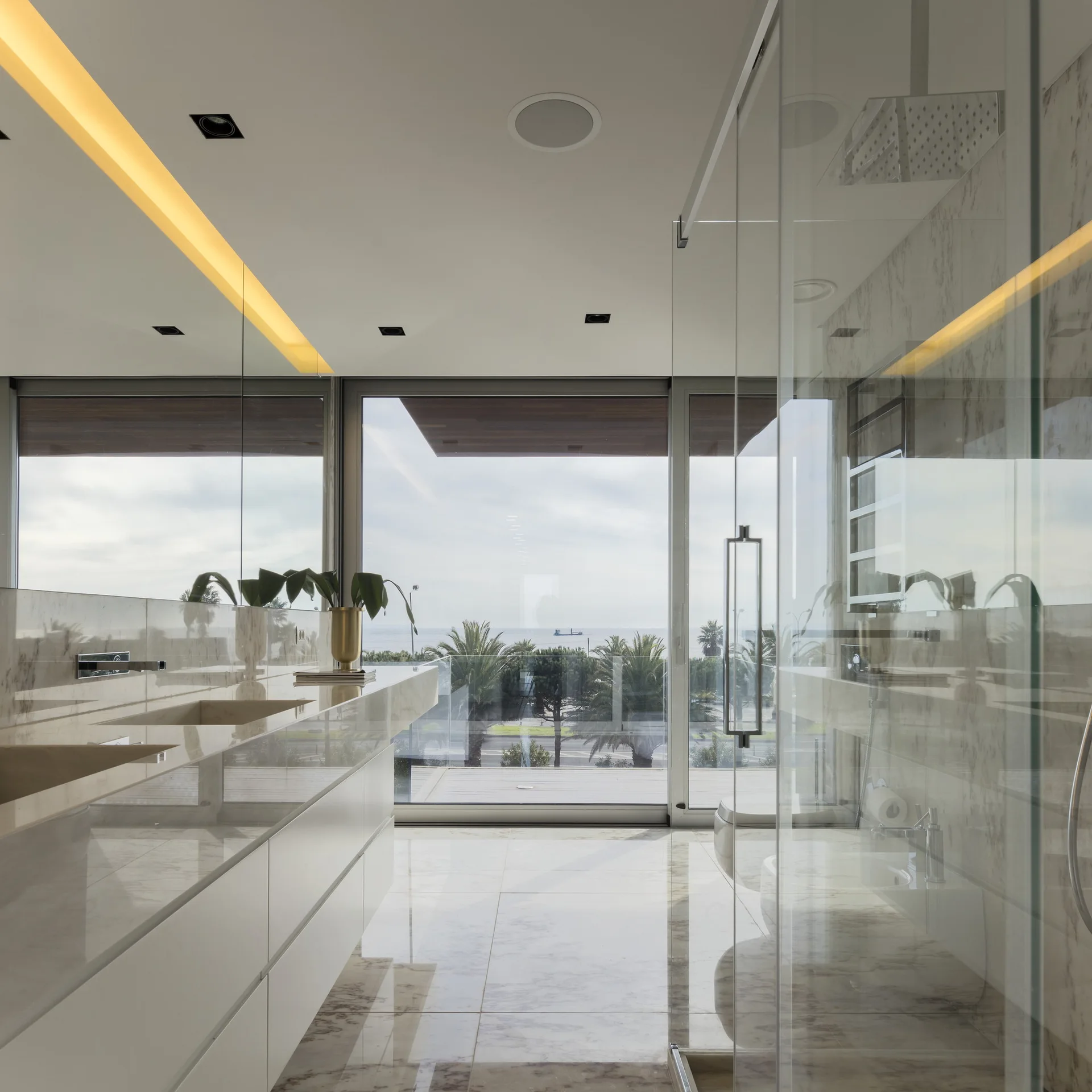 A modern bathroom features a floating marble vanity, polished marble floors, a glass shower, and sliding glass doors opening to an ocean view.