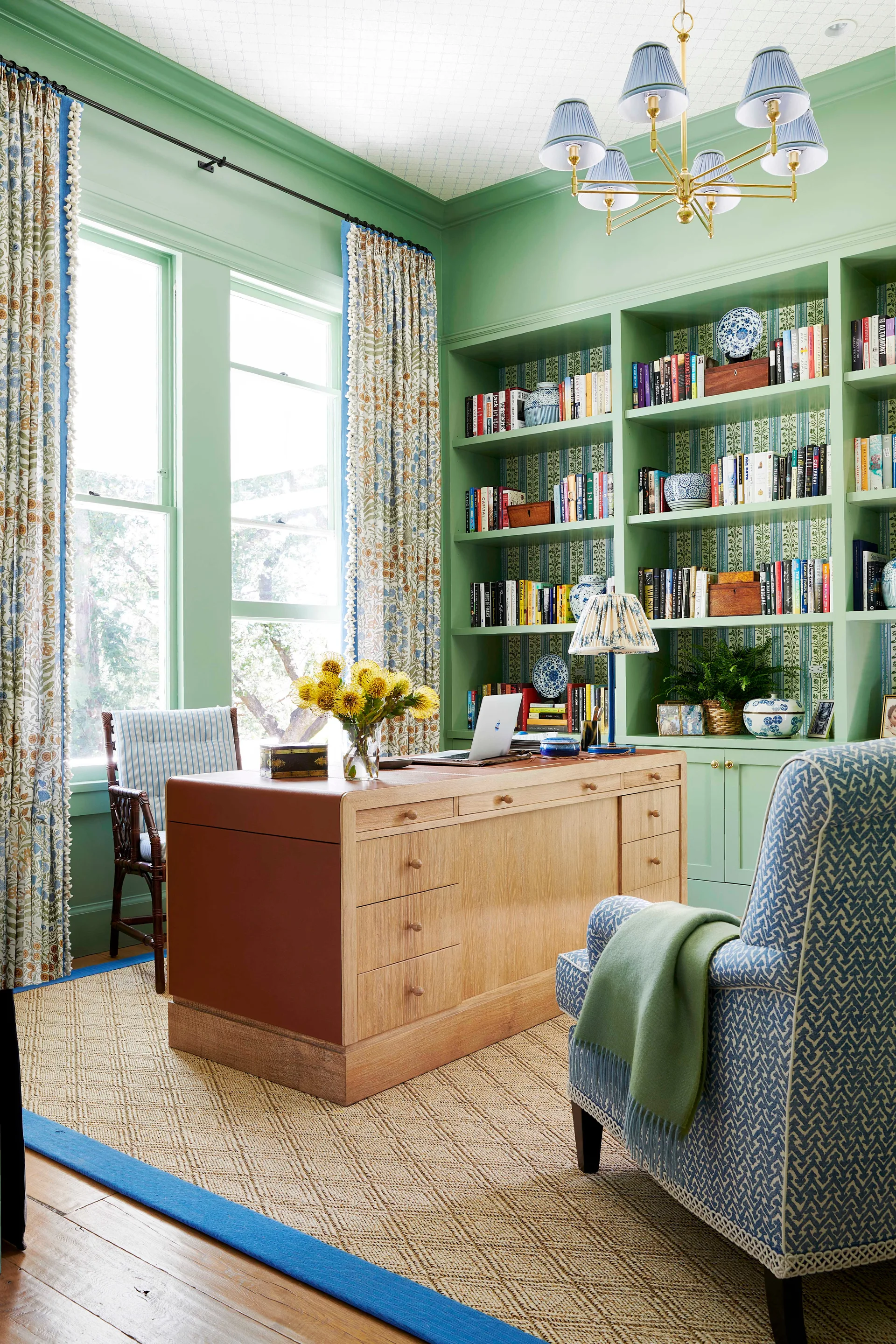 A home office features sage green built-in bookshelves, a light wood desk, a blue patterned armchair, and a textured woven rug.