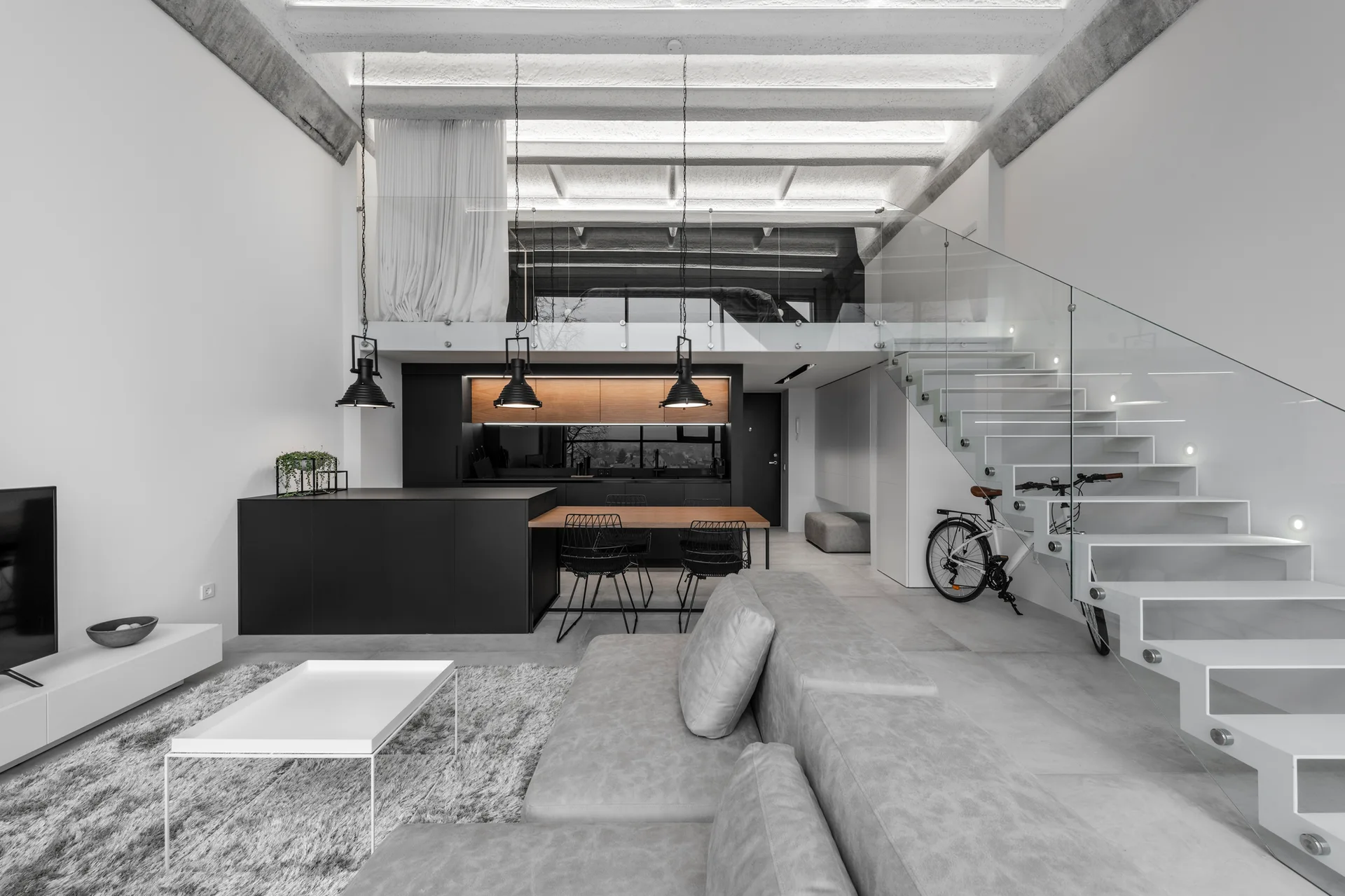 A modern open-plan living room with a grey sectional sofa, black kitchen island, and a white cantilevered staircase with glass railings.