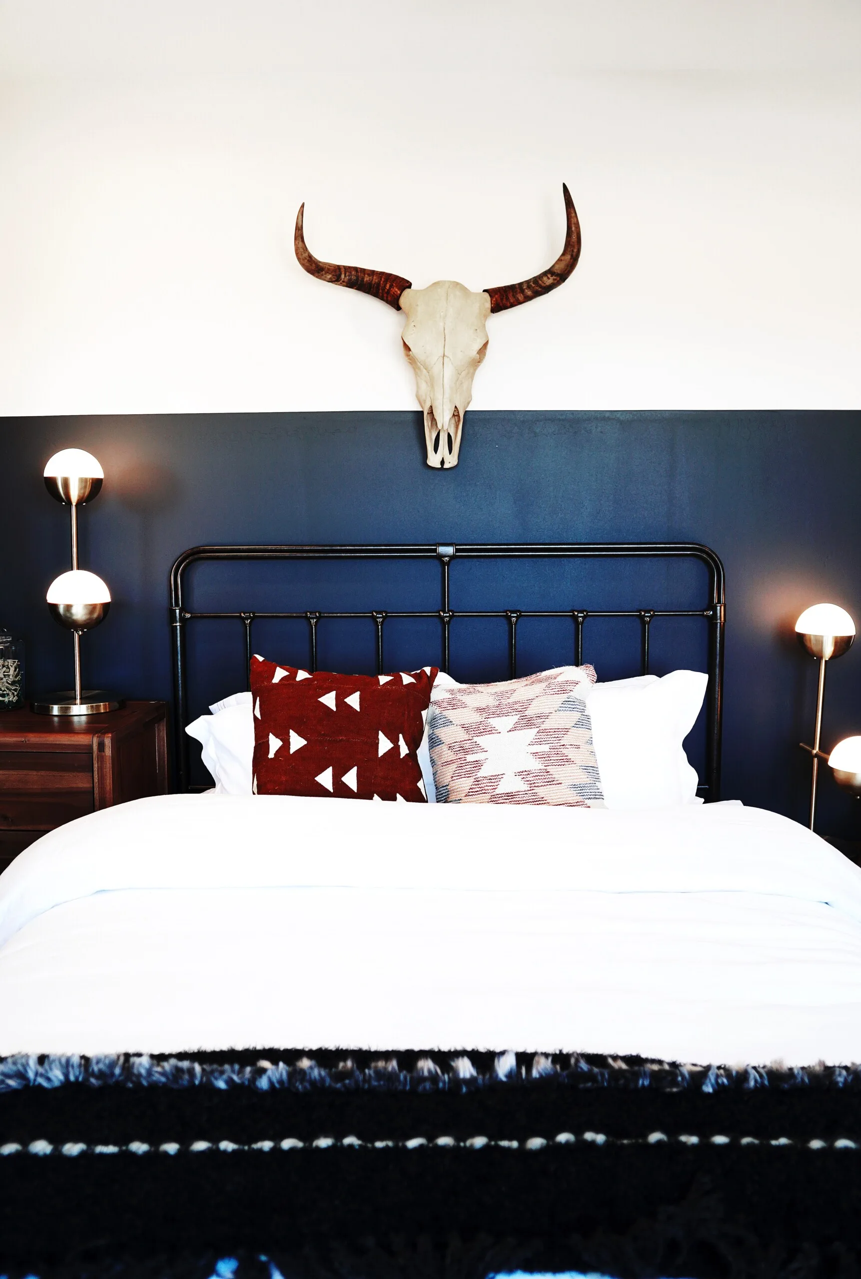 A black metal bed with white bedding and geometric pillows sits against a two-tone navy and white wall topped with a mounted animal skull.