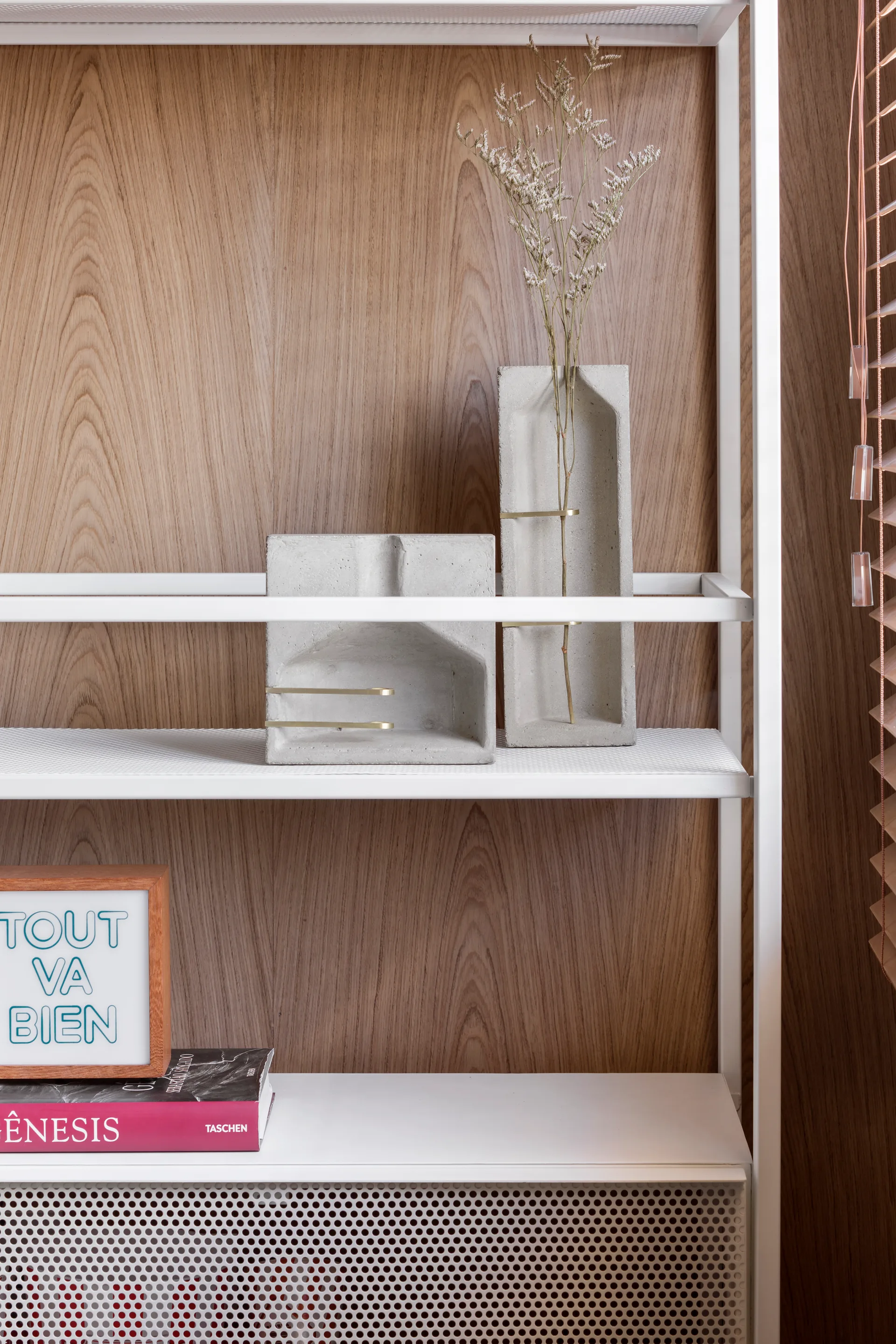 A close-up of a white metal shelf displaying concrete sculptural vases and books against a warm vertical wood grain paneled wall.