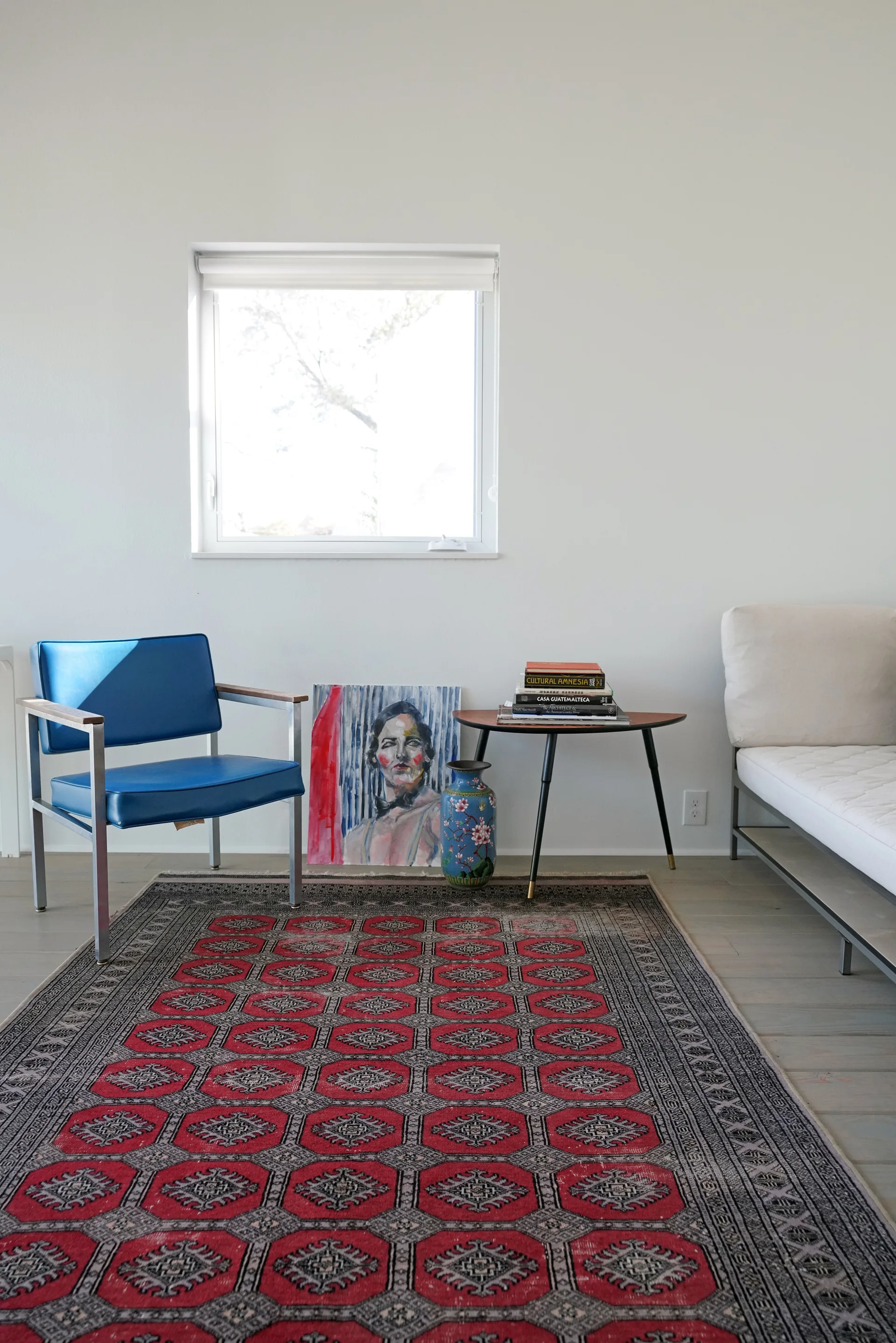 A bright room features a blue metal armchair, a small wood side table, leaning art, and a large red patterned rug on light wood floors.