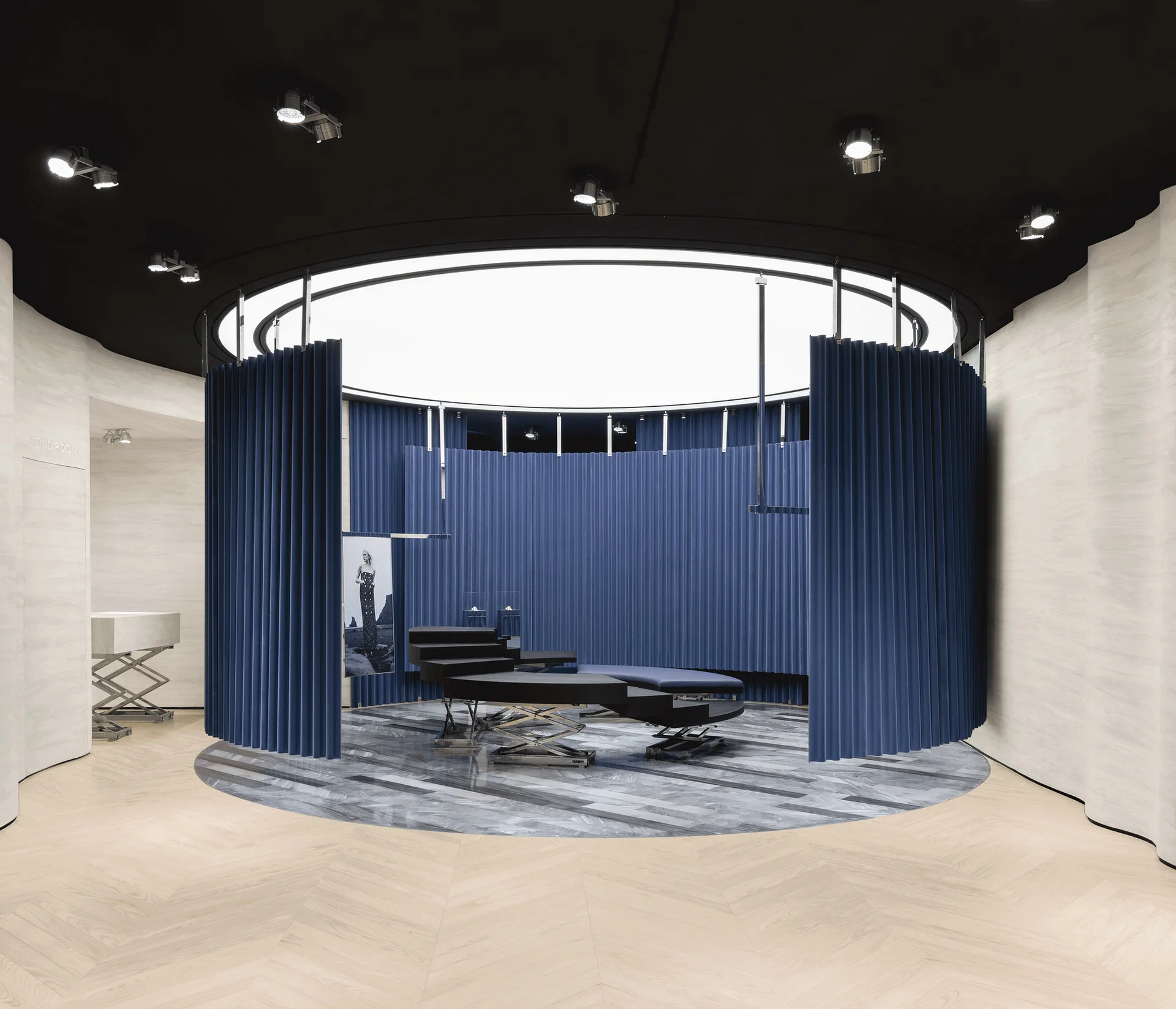 A circular blue pleated curtain surrounds a black tiered display platform set on a grey marble inlay within a light wood chevron floor.