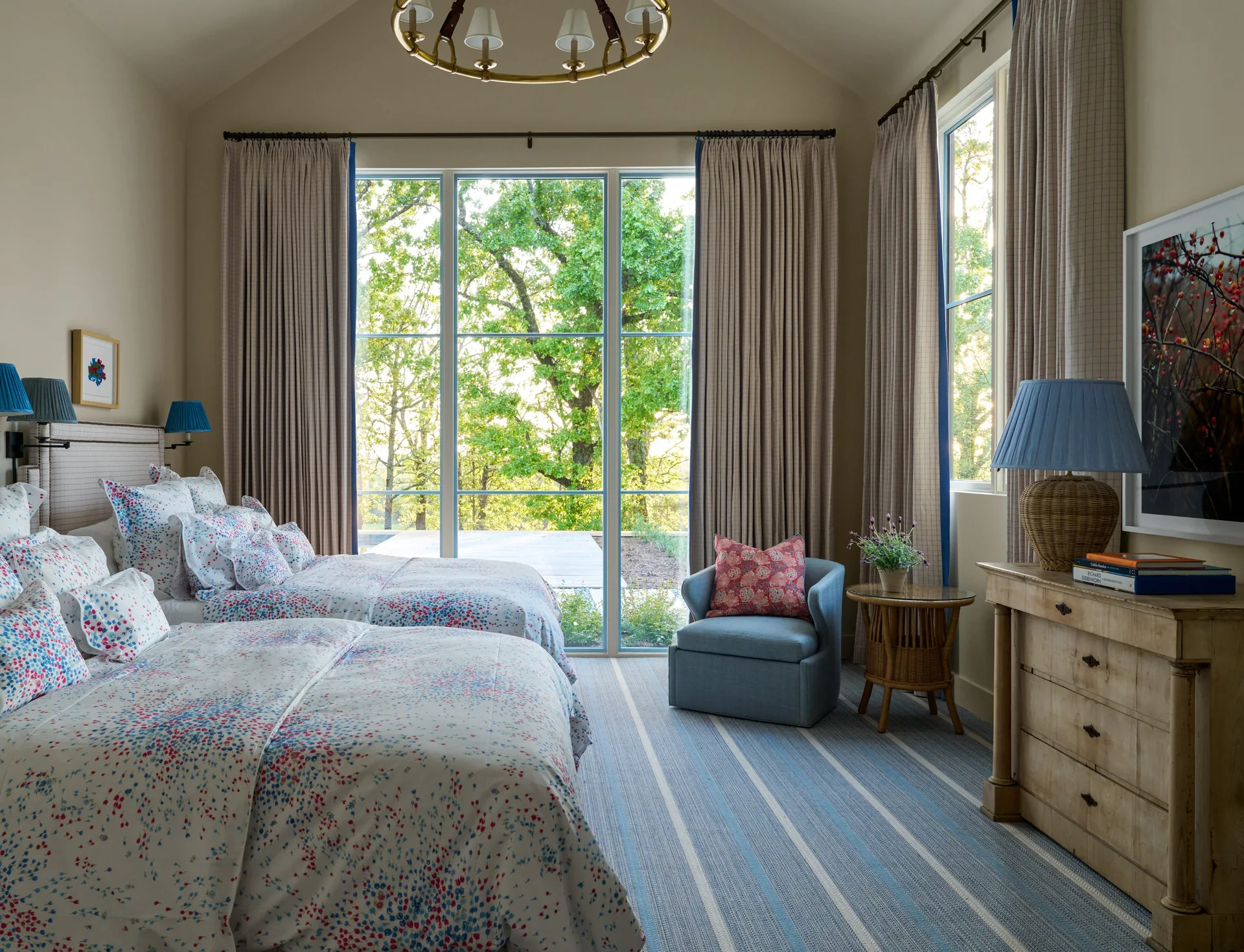 A twin bedroom features two beds with floral bedding, a blue striped rug, an armchair, and a large window looking out onto trees.