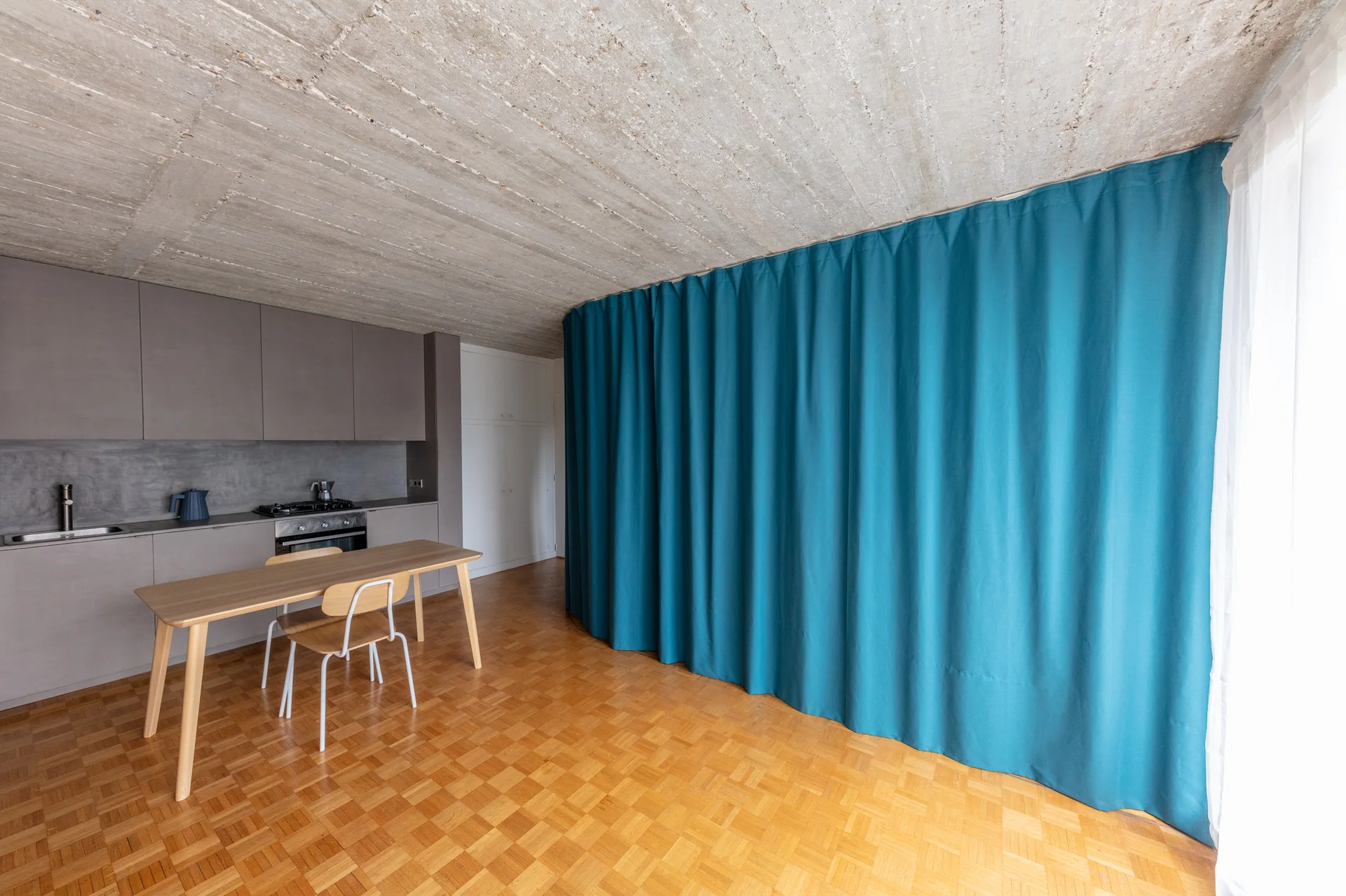 A minimalist kitchen features matte grey cabinets, a wooden dining set, a raw concrete ceiling, and a large curved blue curtain used as a space divider.