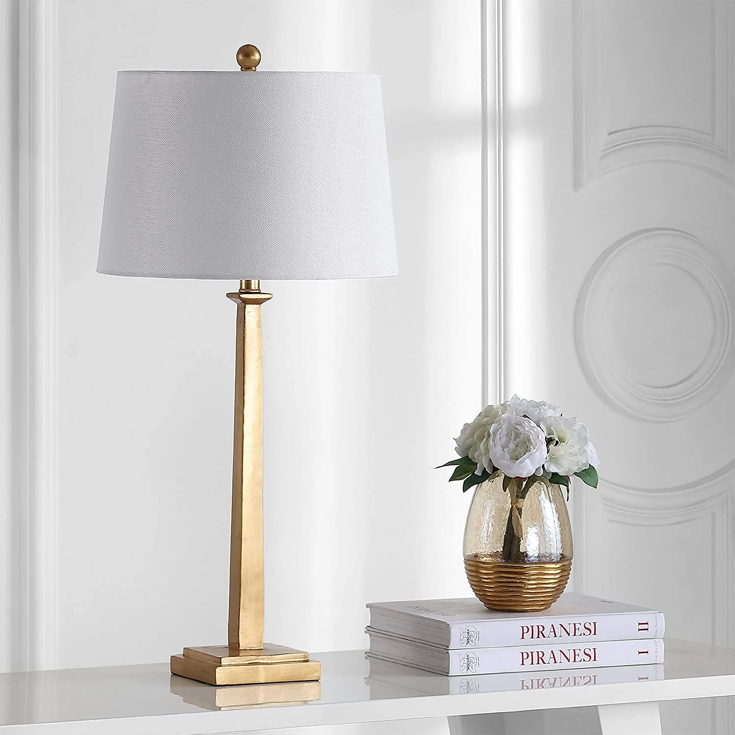 A tall gold column table lamp with a white drum shade sits on a white table next to stacked books and a small flower vase.