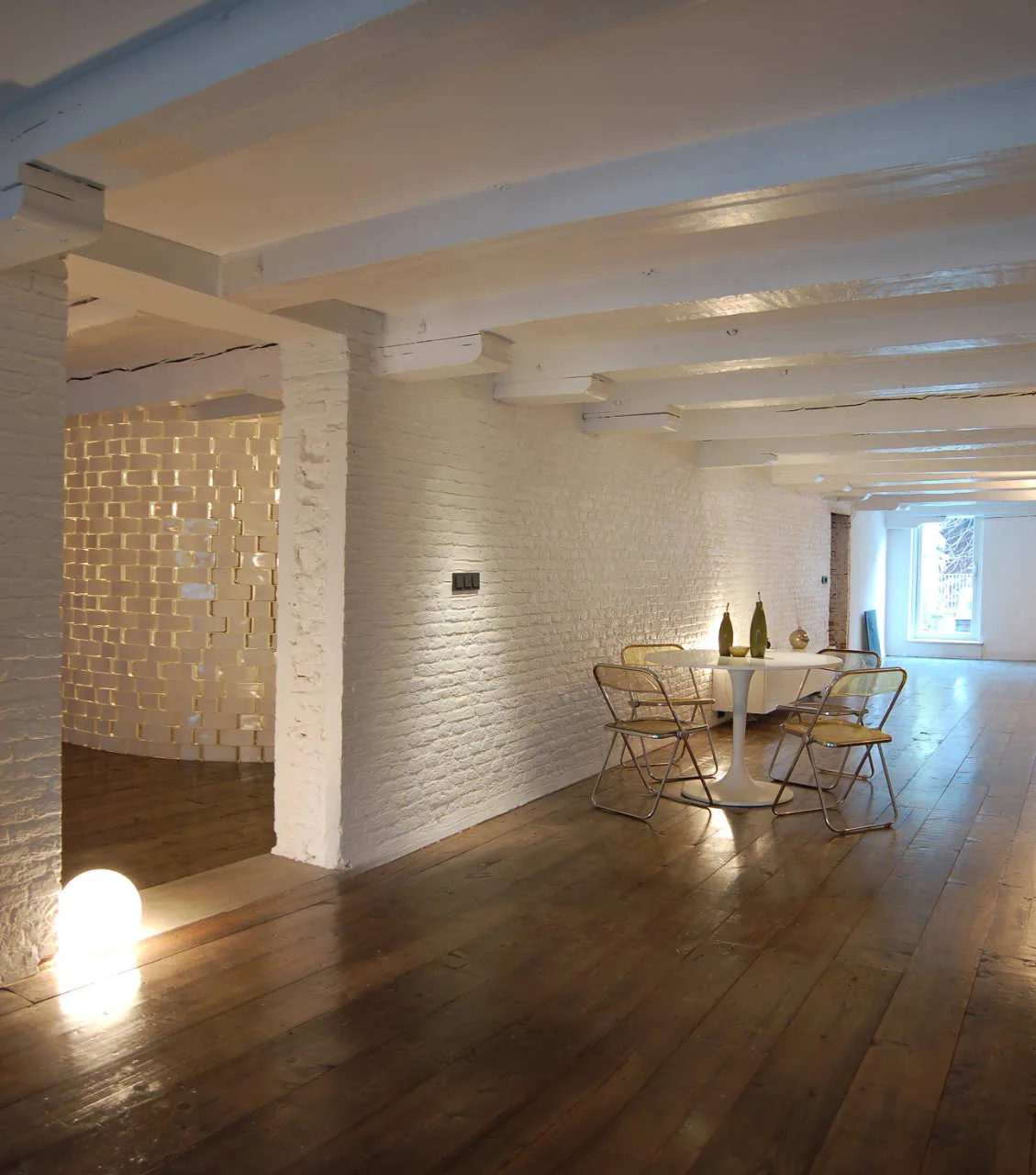 An open dining area features a white tulip table with metal and cane chairs, set against a painted white brick wall and dark wood floors.