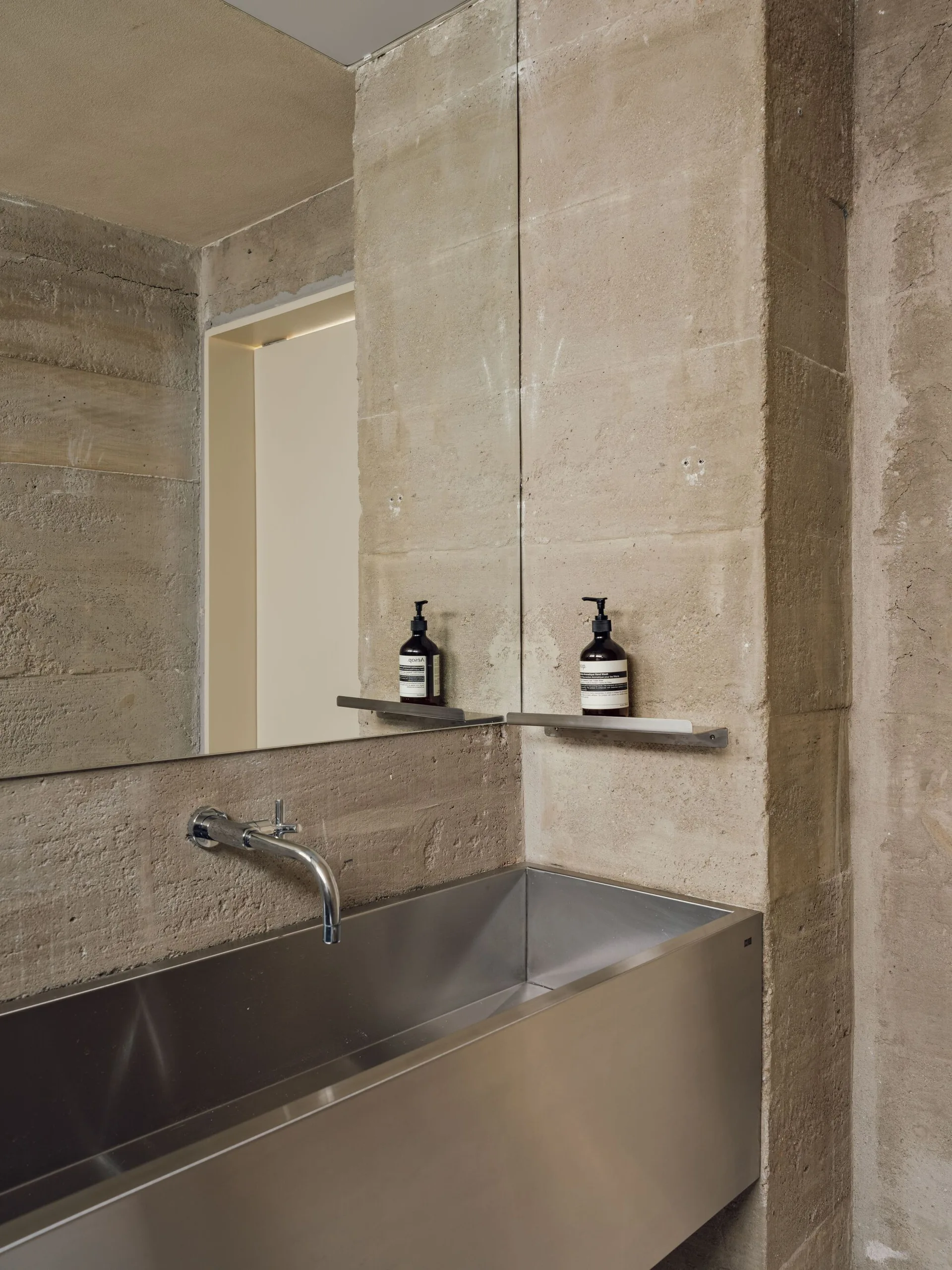 A stainless steel trough sink is mounted against a textured concrete wall under a large rectangular mirror in a minimalist bathroom.