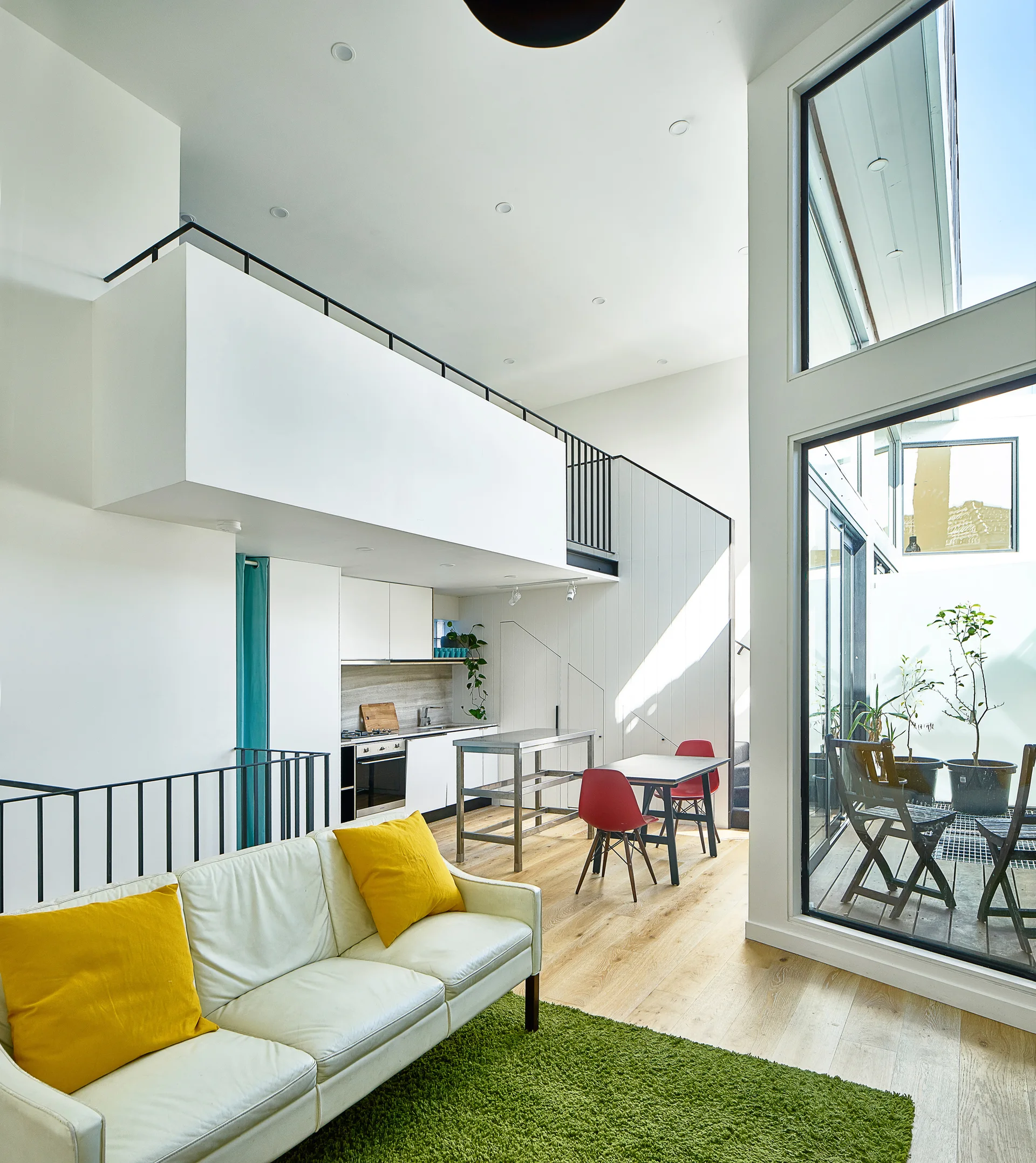A modern loft apartment living room with a white sofa on a green rug, a dining area, and a kitchen under a mezzanine balcony.