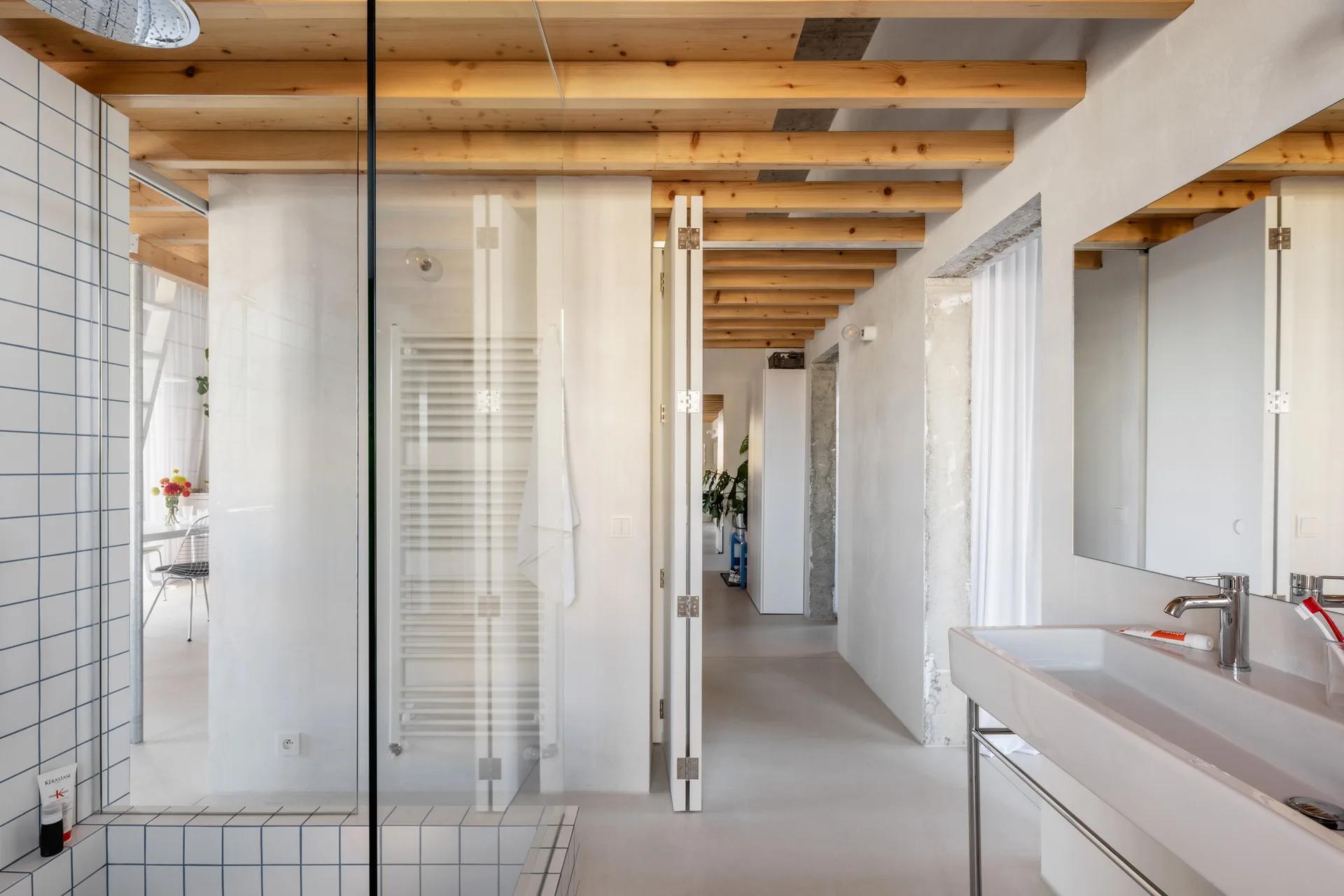 A minimalist bathroom featuring exposed timber ceiling beams, a glass shower partition with grid tiling, and a modern wall-mounted sink vanity.