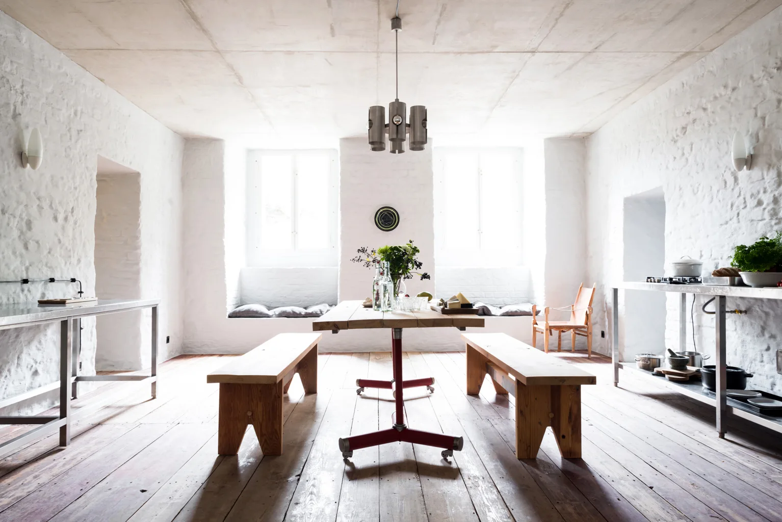 A rustic dining room featuring a wooden table with red metal legs and matching wood benches set between deep window seats against textured white brick walls.