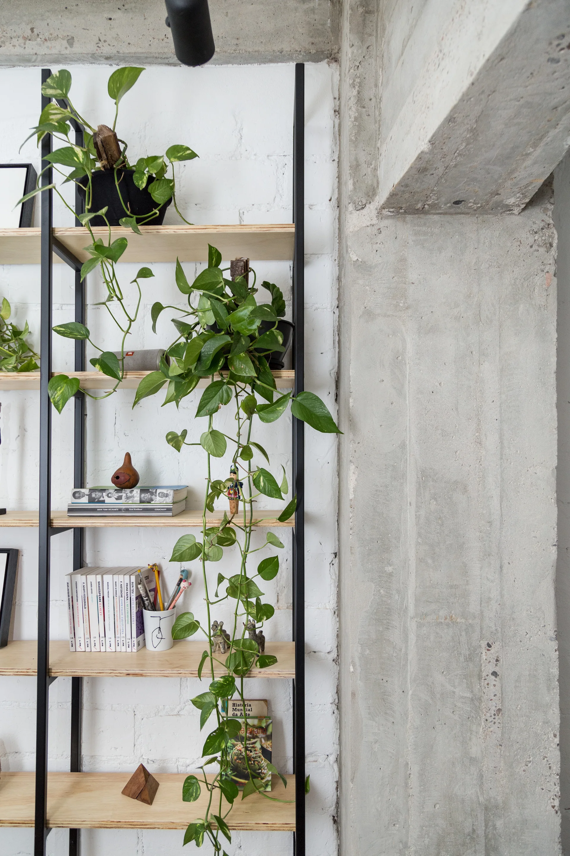 An open shelving unit with light wood shelves and a black metal frame stands against a white brick wall next to a raw concrete pillar.
