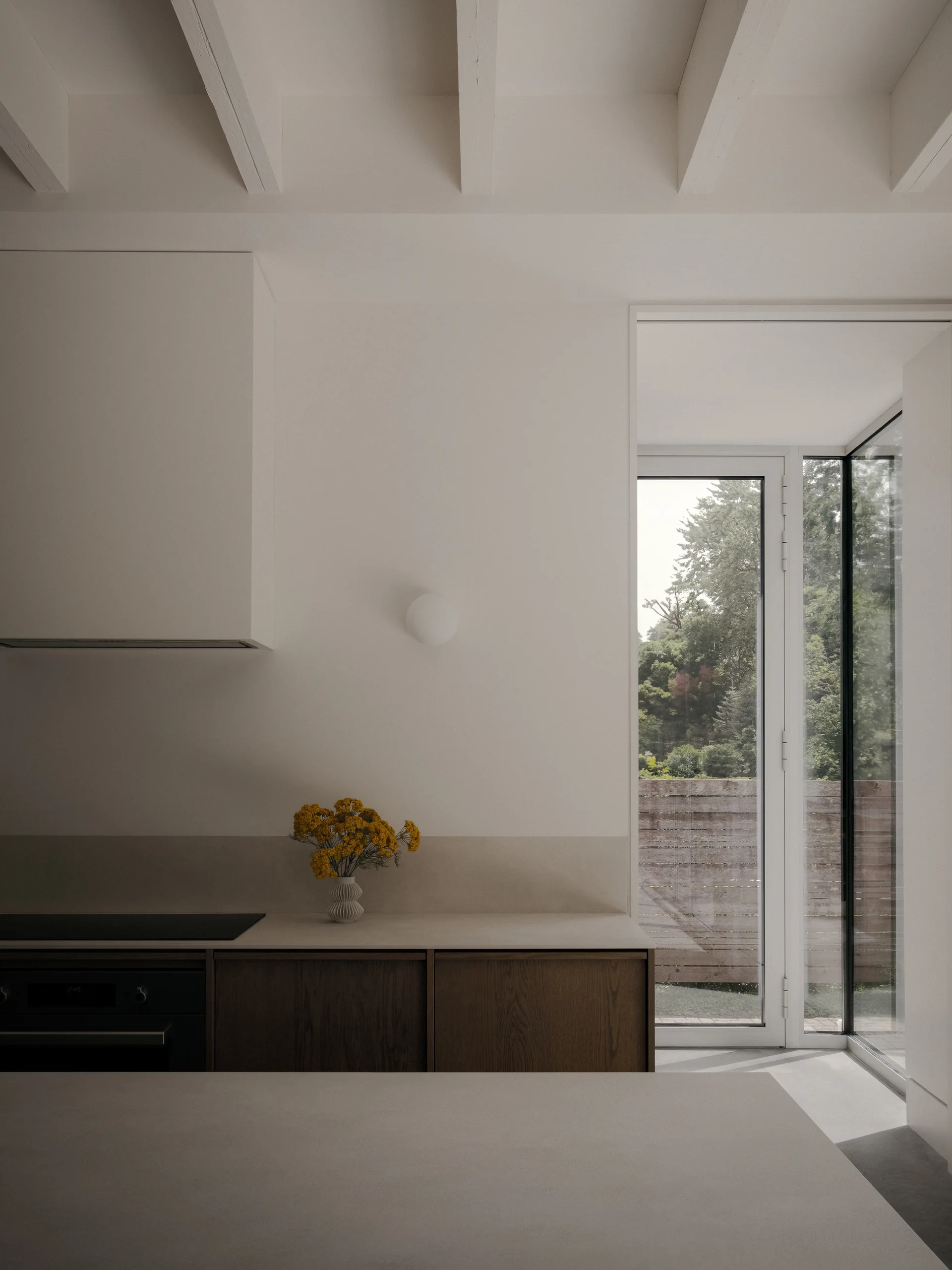 A minimalist kitchen features warm wood lower cabinets, a white countertop, painted ceiling beams, and a tall glass door opening to the outdoors.