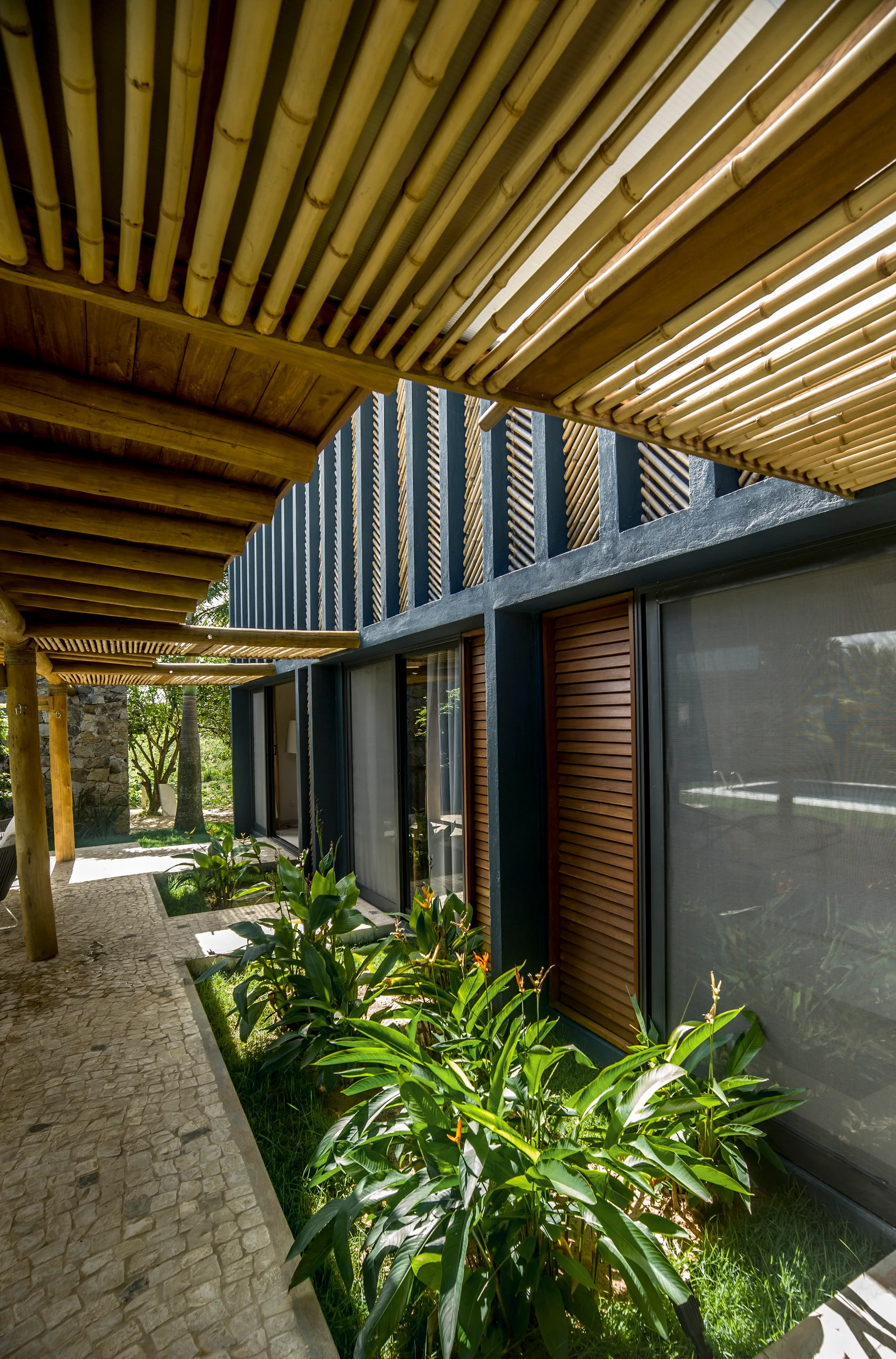 A stone paved pathway runs alongside a dark gray building facade with large glass doors, wooden shutters, and a slatted bamboo pergola overhead.
