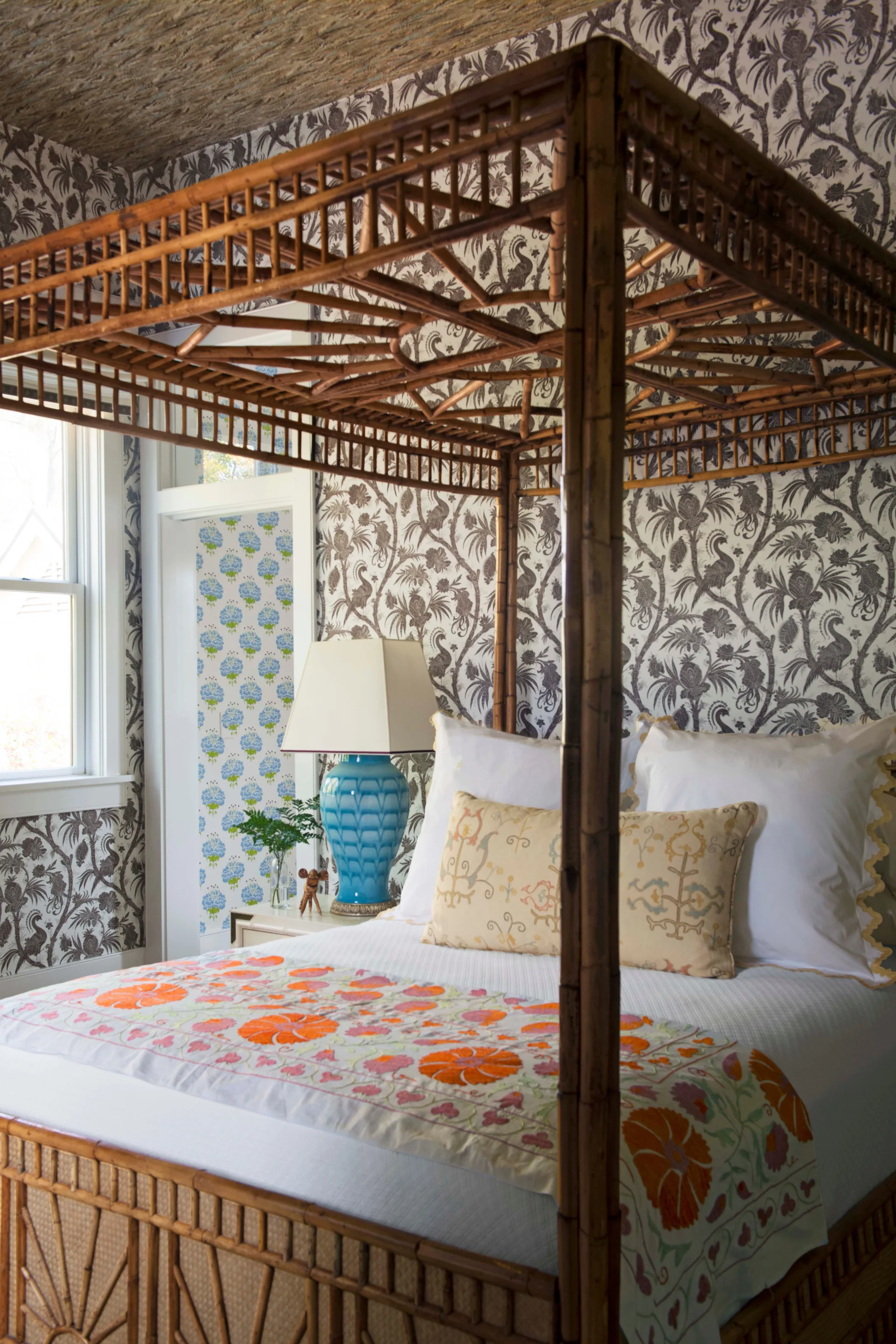 A bedroom features a bamboo canopy bed, grey and white botanical wallpaper, and a blue ceramic table lamp.