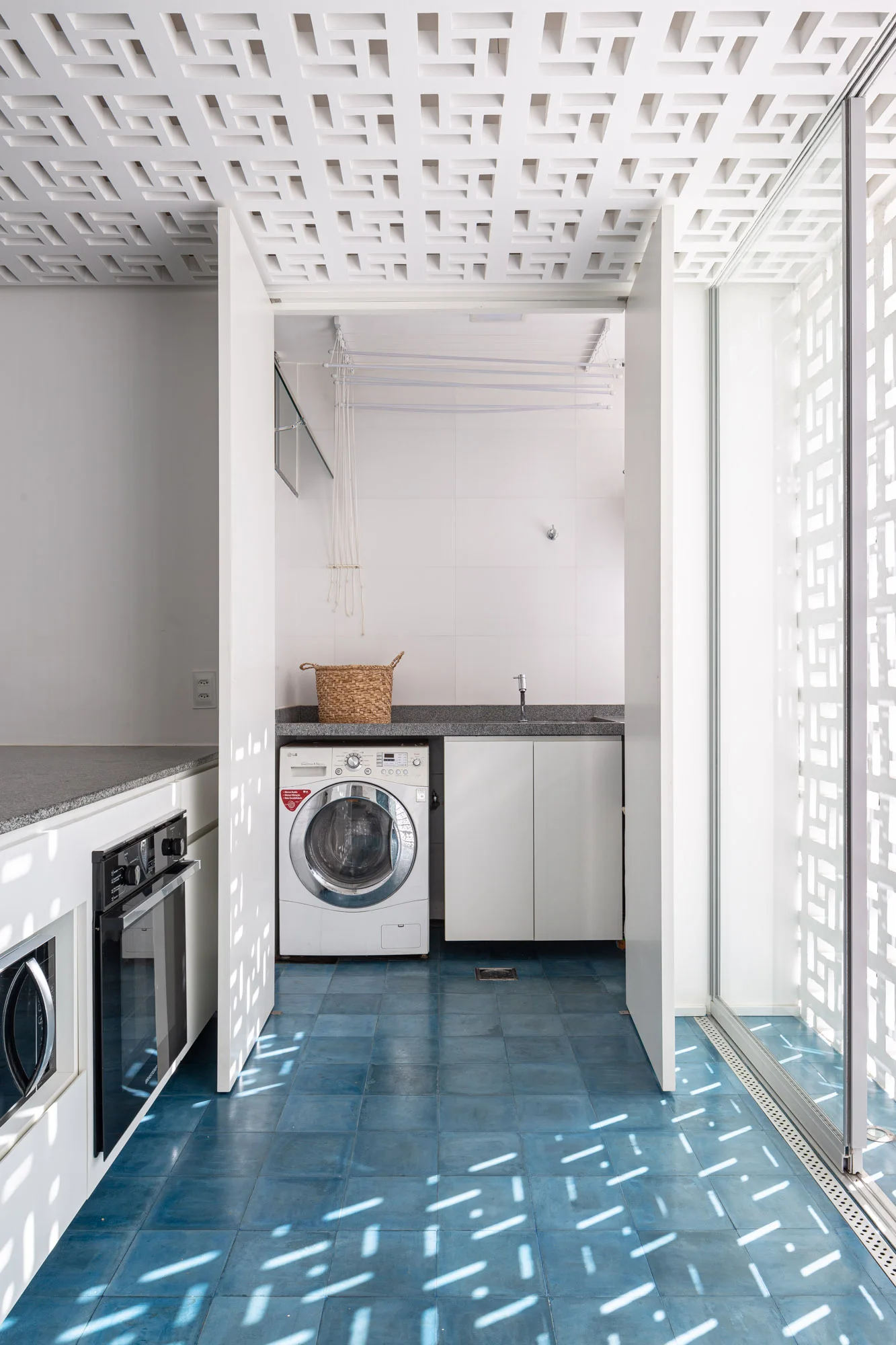 A bright modern laundry room featuring blue square tiled floors, a white washing machine, and a geometric lattice ceiling screen.