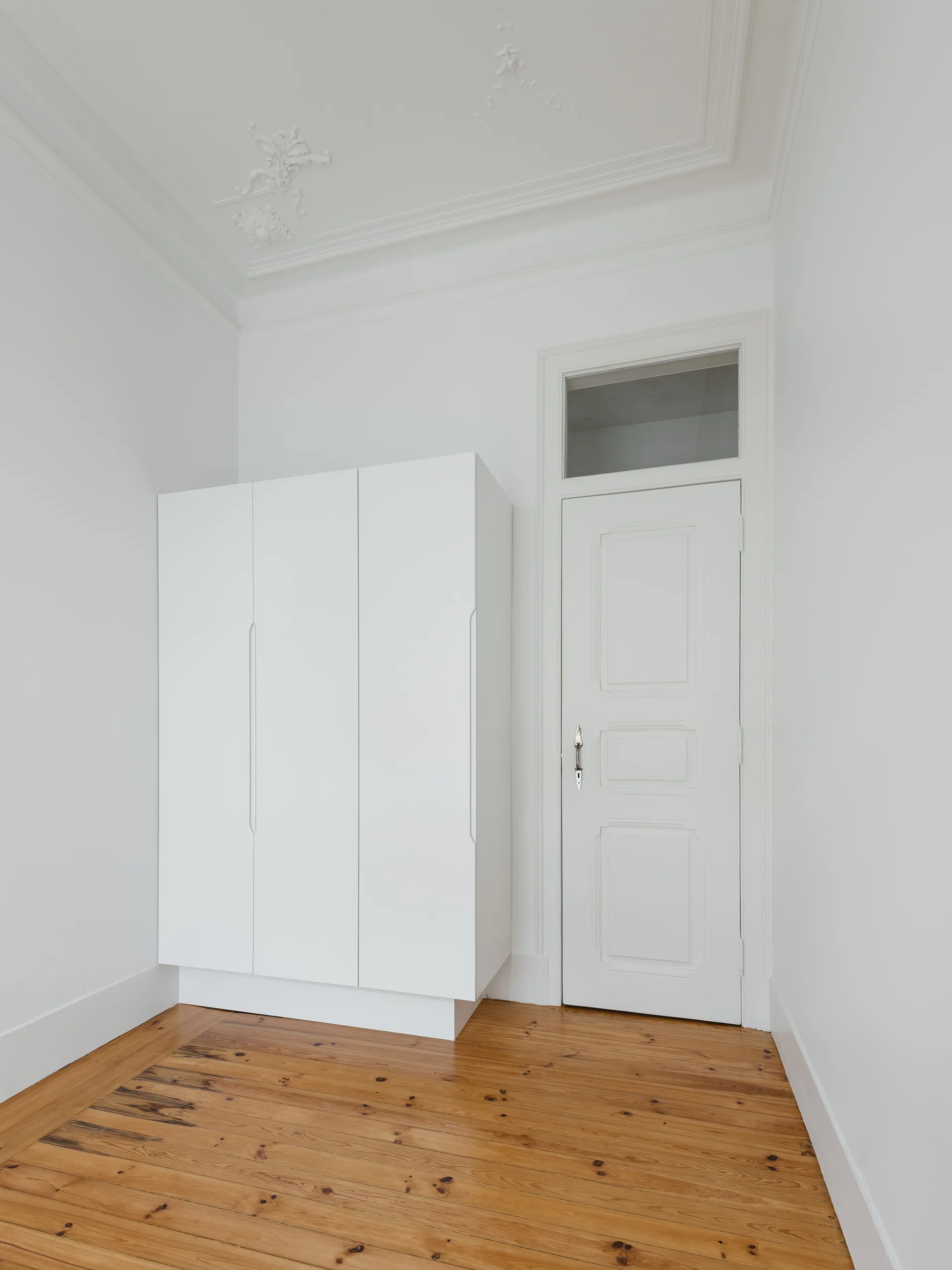A matte white modern wardrobe stands on natural wood floors next to a traditional paneled door with a transom window under an ornate ceiling.