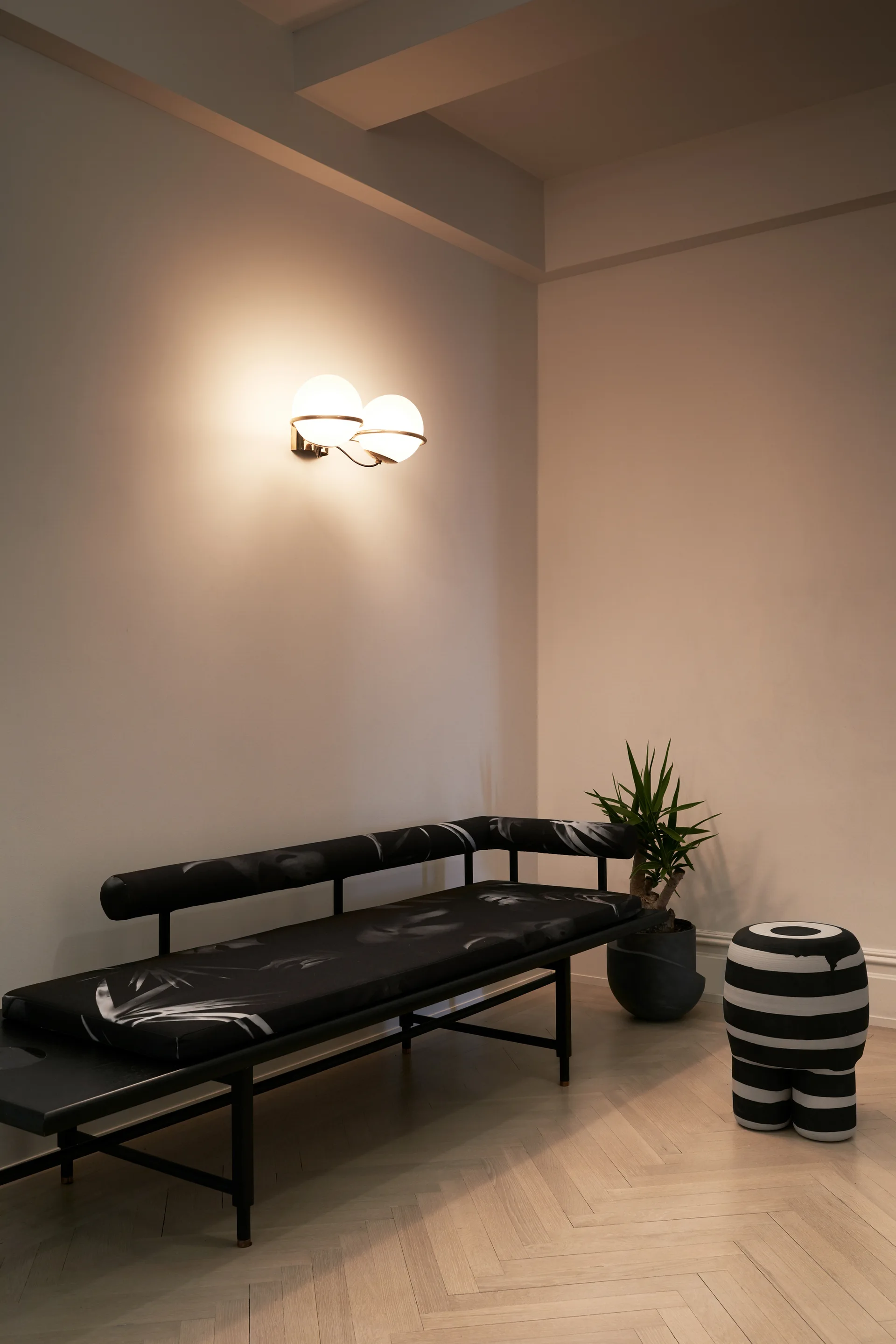 A black patterned bench sits below a dual globe wall sconce in a minimalist corner with a striped stool and herringbone wood flooring.