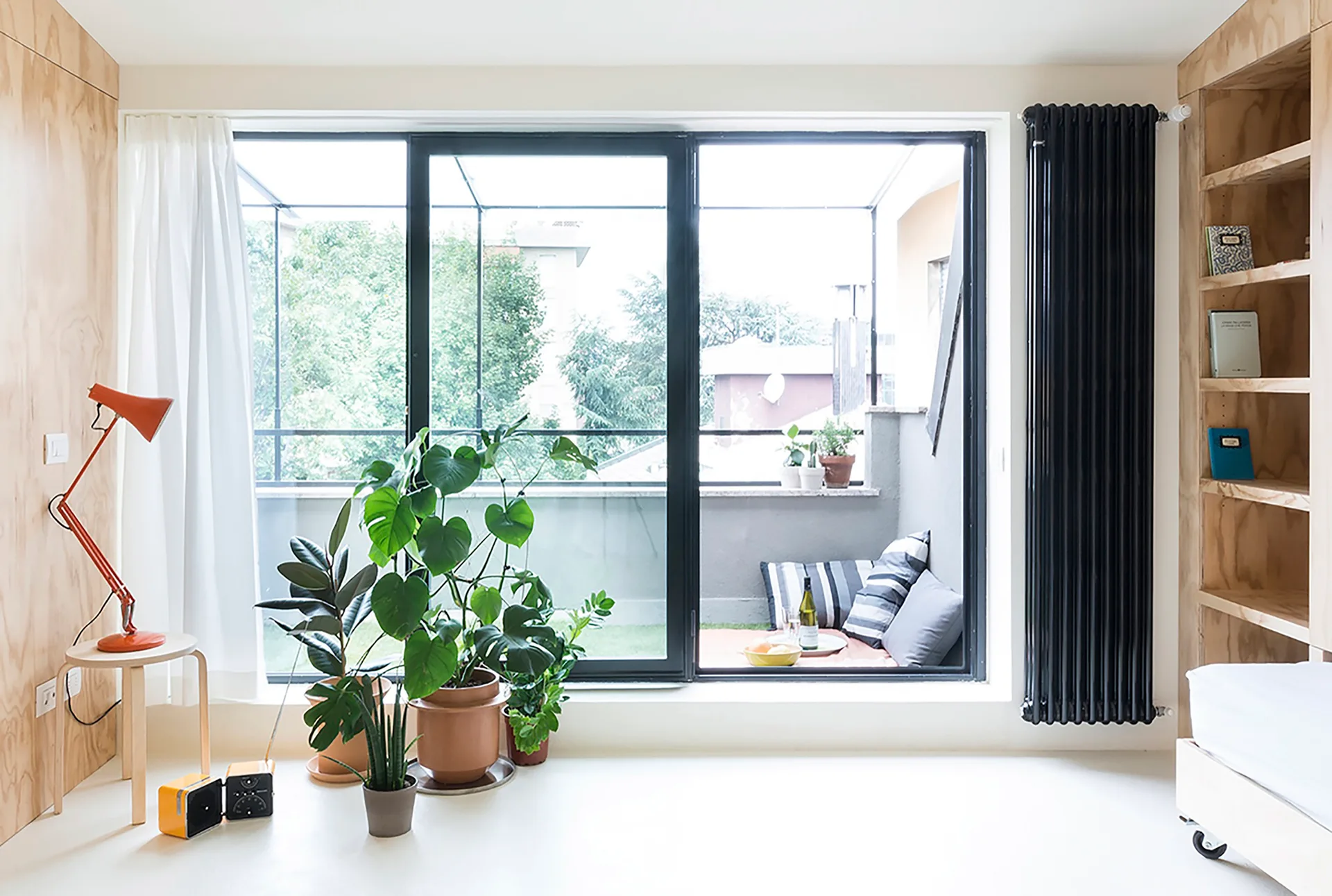 A bright room with light plywood walls, built-in shelving, a tall black column radiator, and sliding glass doors opening onto a balcony.