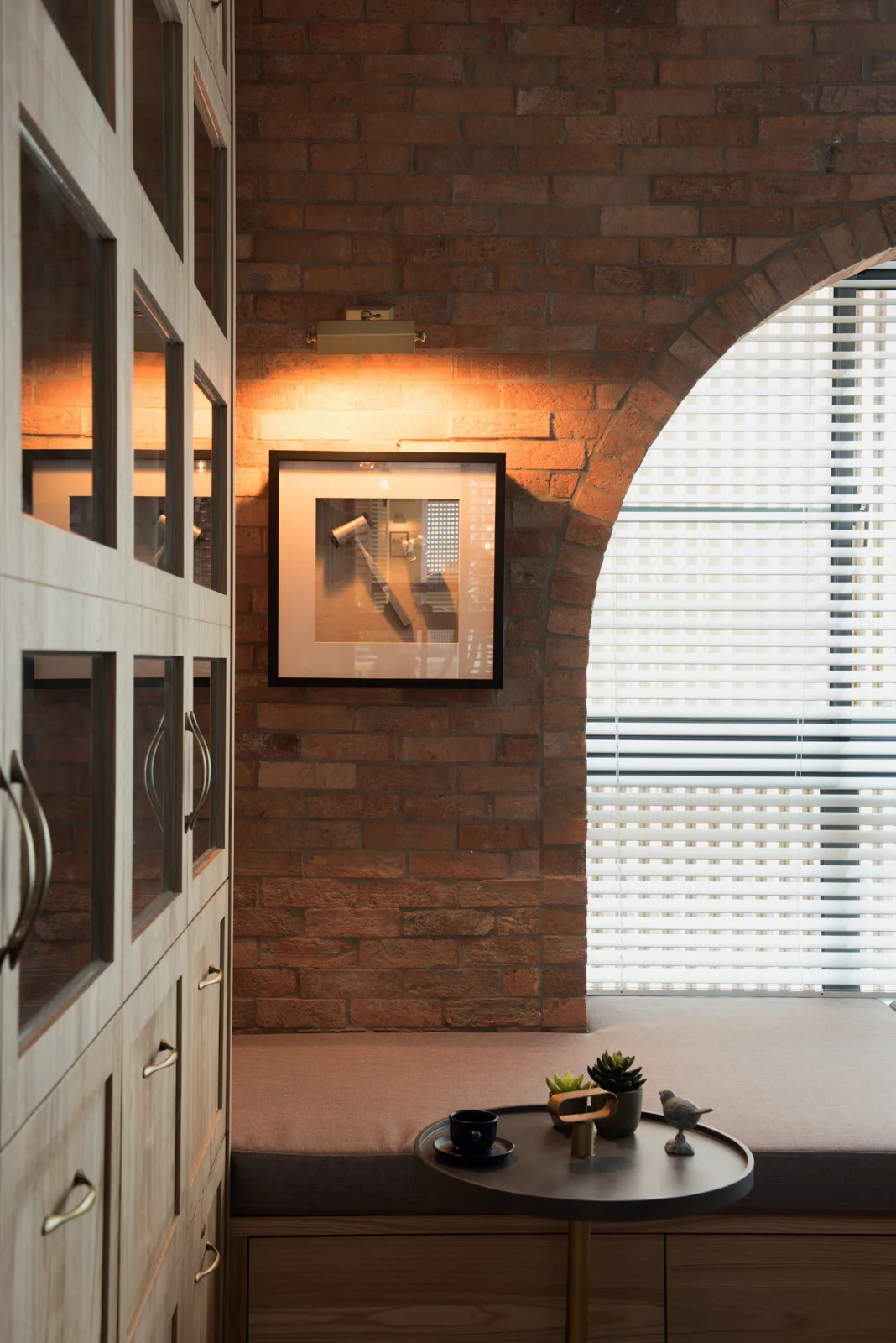 A built-in bench seat sits next to a light wood cabinet under an exposed brick arch with venetian blinds.