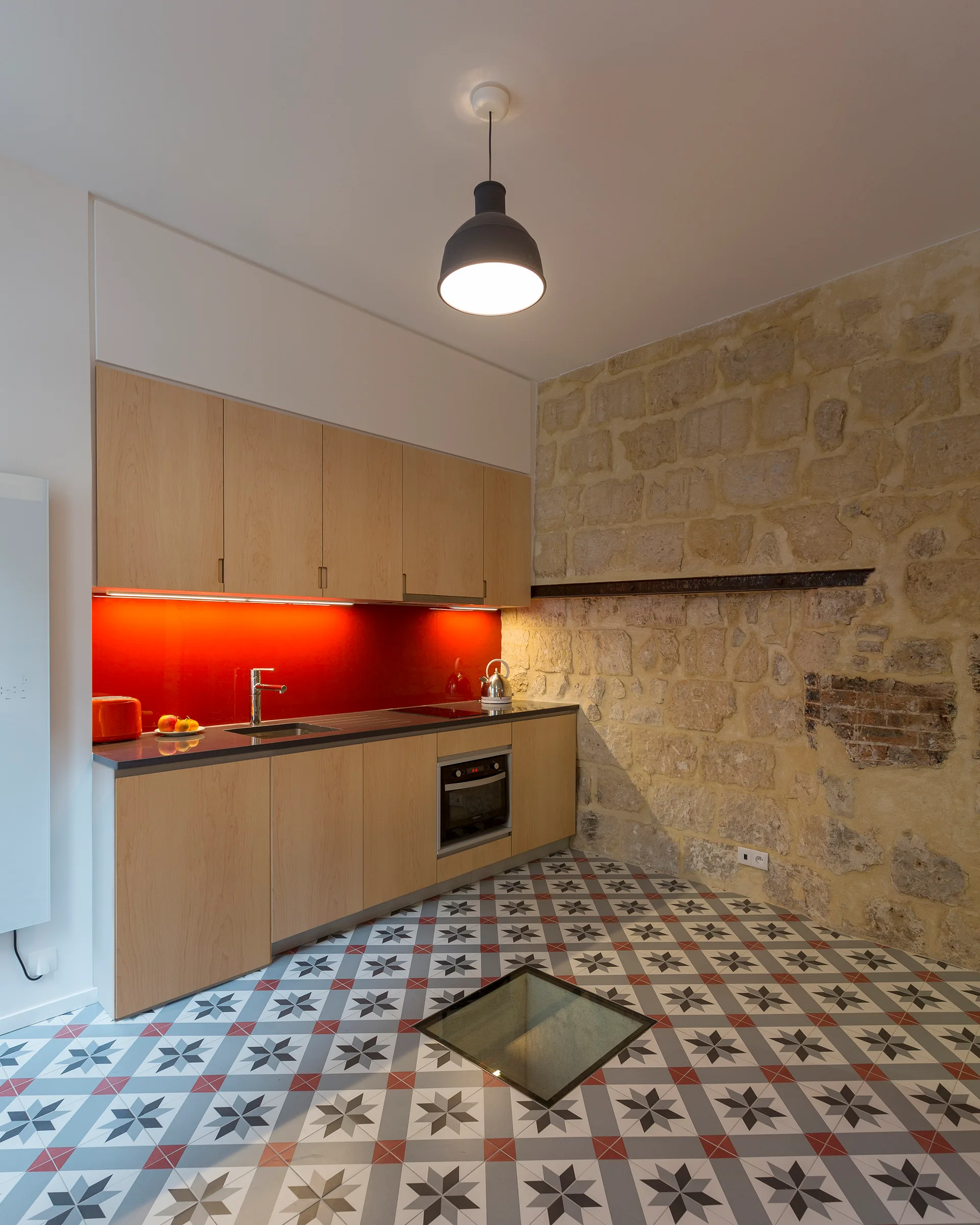 A modern kitchen featuring light wood cabinets, a vibrant red backsplash, an exposed stone wall, and geometric patterned floor tiles with a glass insert.