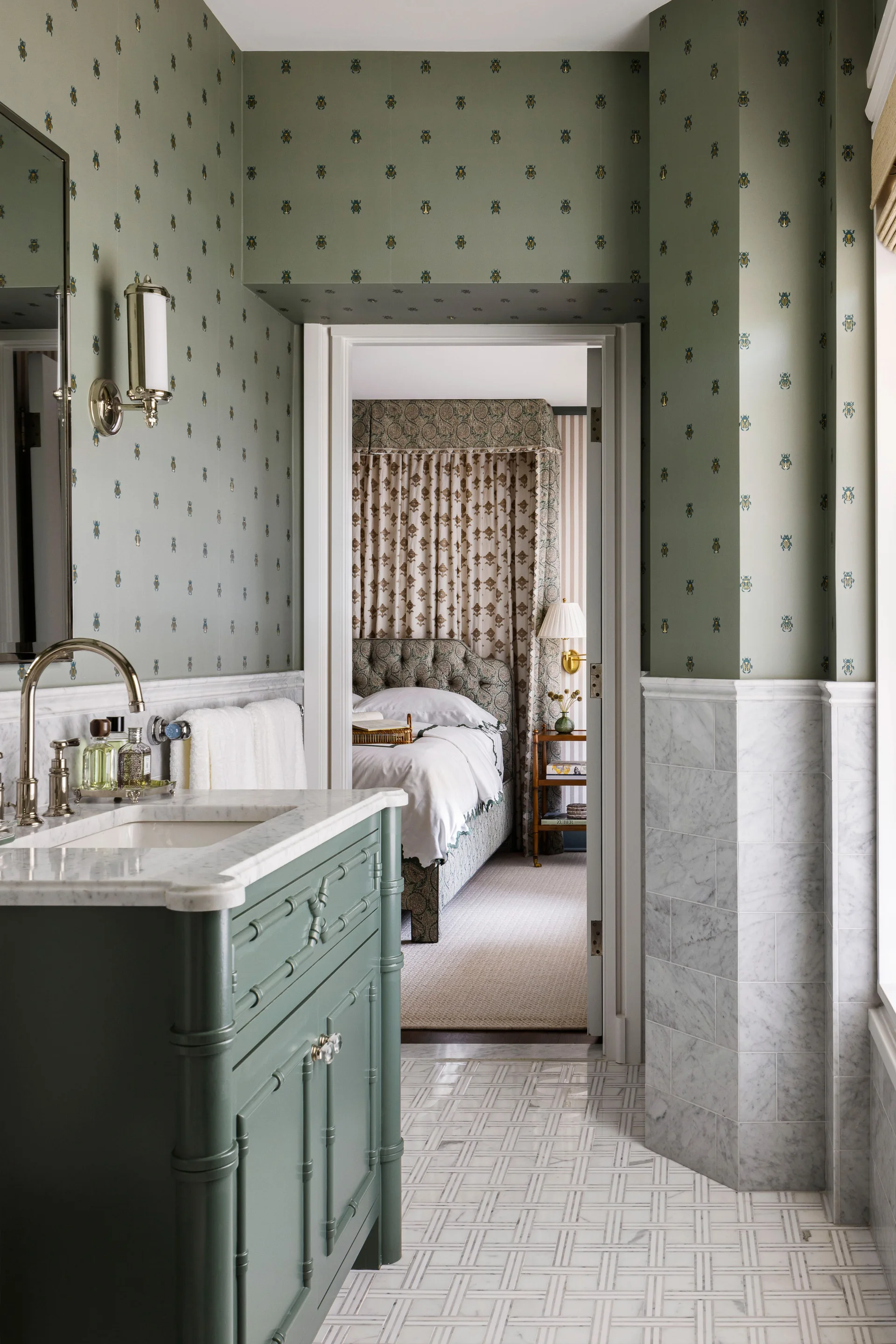 A green bathroom with patterned wallpaper, marble tile wainscoting, and a green vanity with a marble countertop, showing a bedroom doorway beyond.