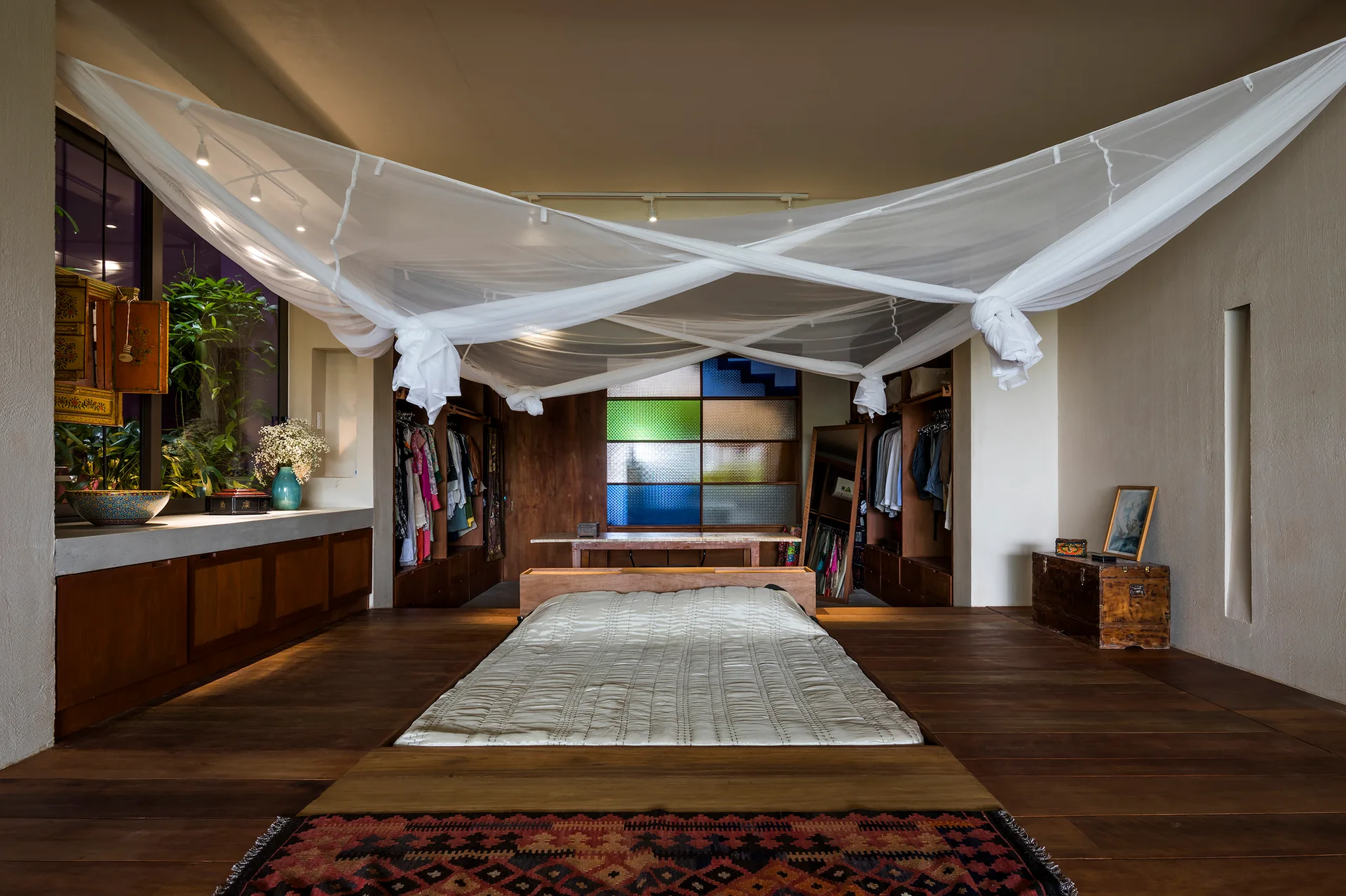 A low platform bed with white canopy netting sits on dark wood floors in a rustic bedroom featuring built-in closets and a stained-glass panel.