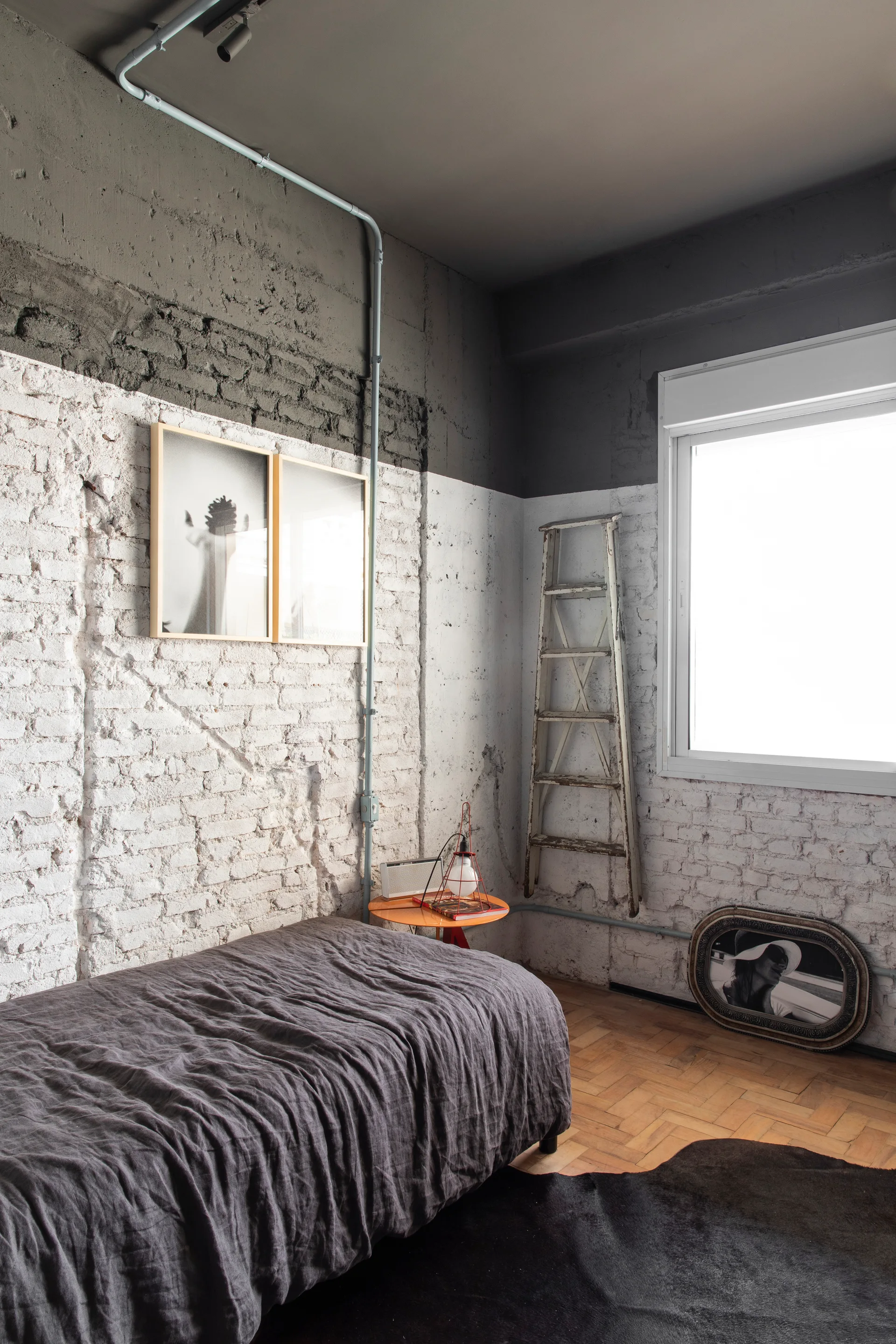 An industrial bedroom features a dark gray bed, painted white brick walls with a dark gray upper border, parquet flooring, and a decorative wooden ladder.