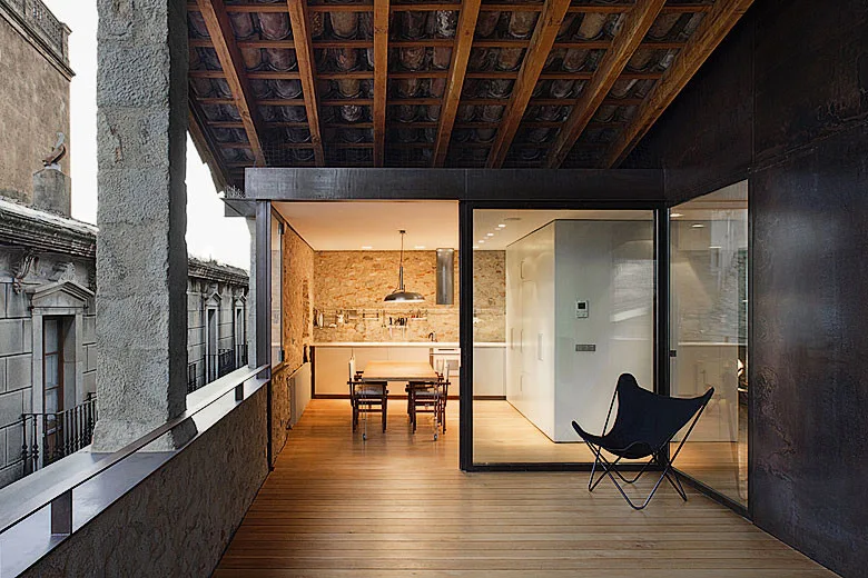 A wooden balcony terrace with a black butterfly chair and an exposed timber ceiling connects to an indoor kitchen with an exposed stone wall.