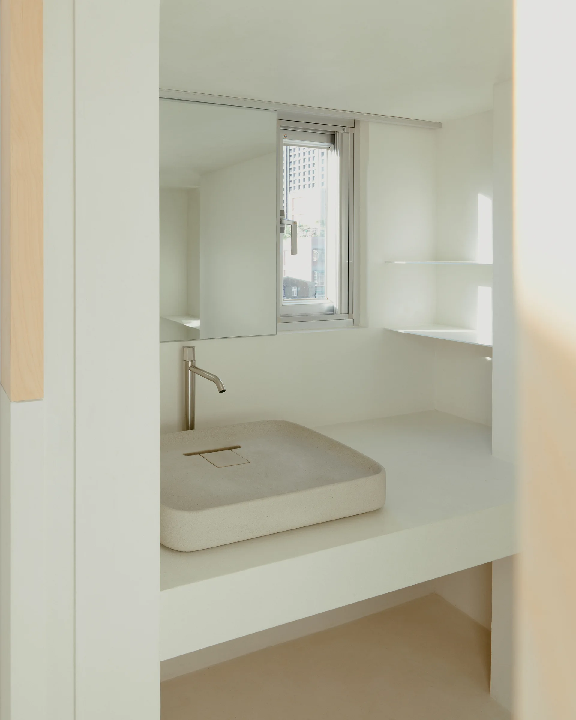 A minimalist bathroom vanity featuring a rectangular stone vessel sink, a wall-mounted silver faucet, a frameless mirror, and white open shelves.