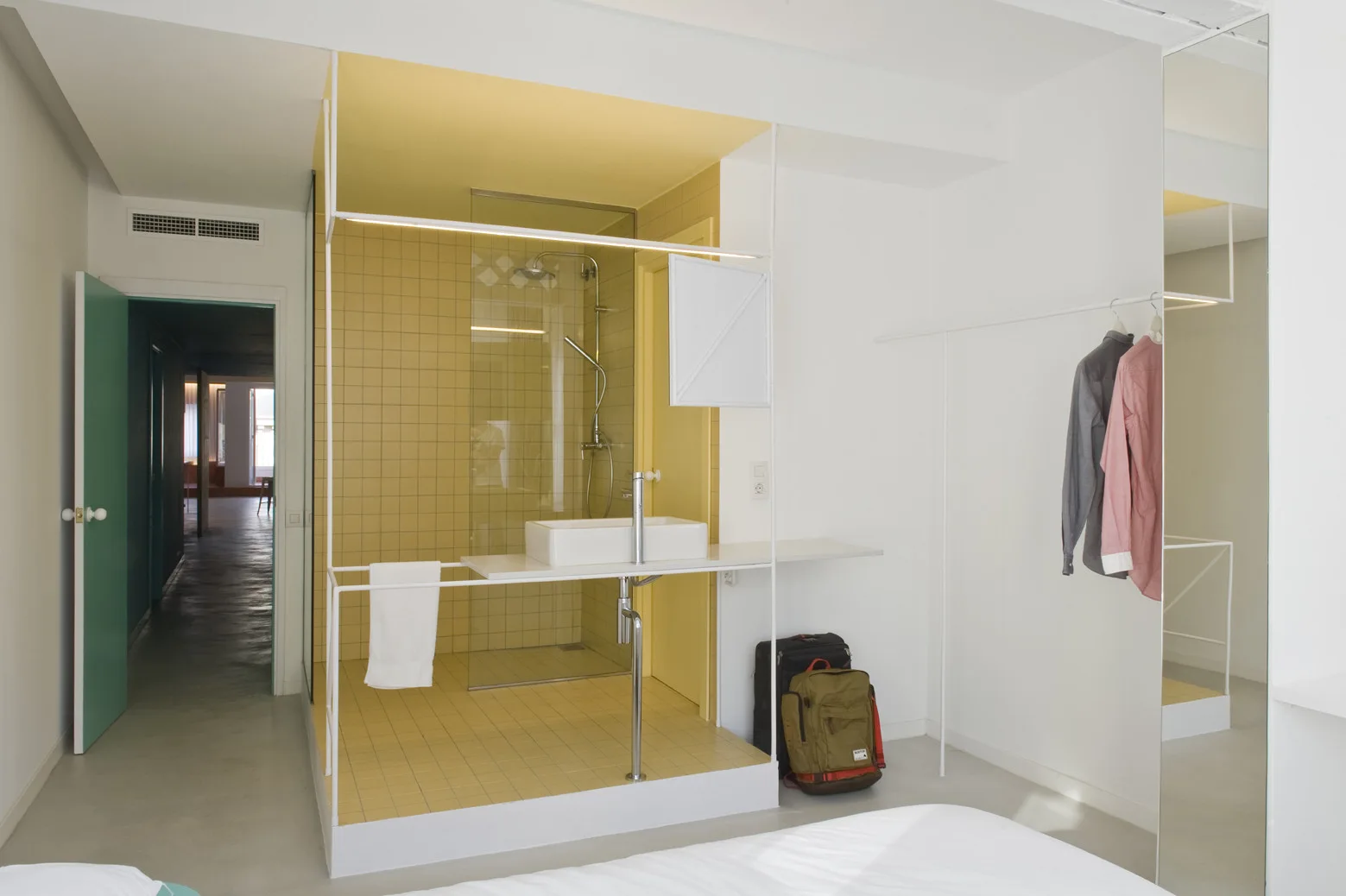 A modern white bedroom featuring a raised glass-enclosed bathroom pod lined with bright yellow grid tiles and a white rectangular sink.