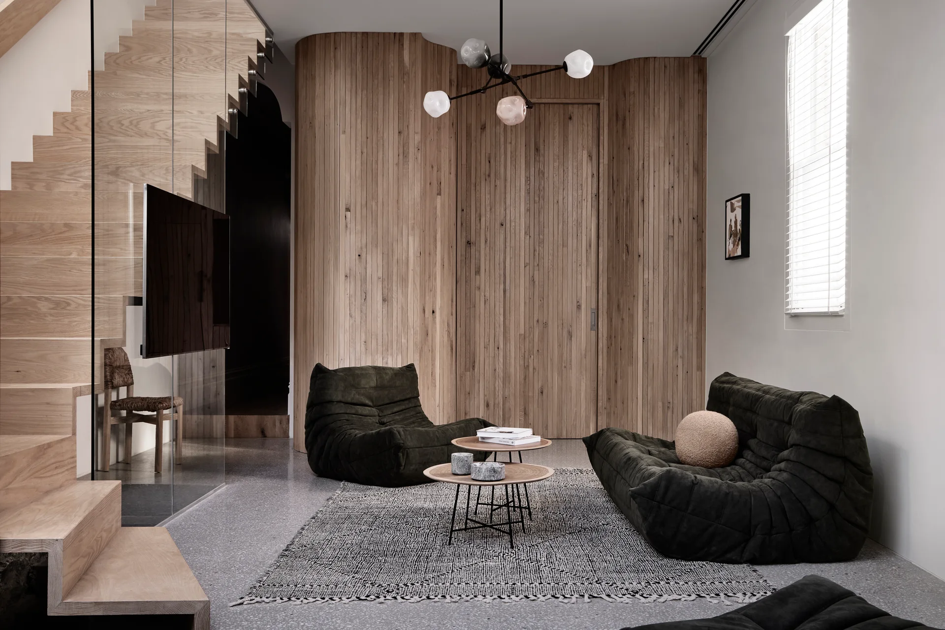 A contemporary living area features dark green quilted chairs, a curved vertical wood slat wall, and a wooden staircase.