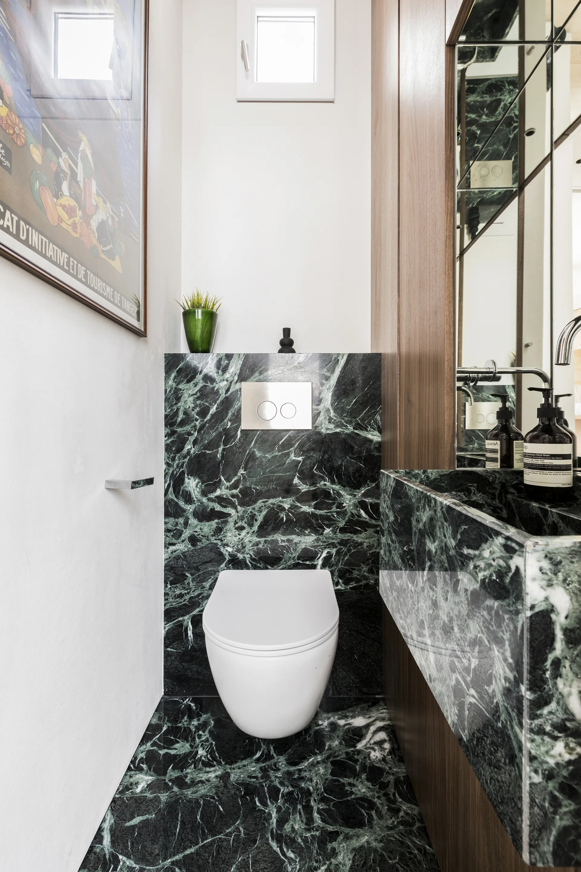 A modern powder room with a white wall-mounted toilet, dark green veined marble floors, and warm wood vanity paneling.