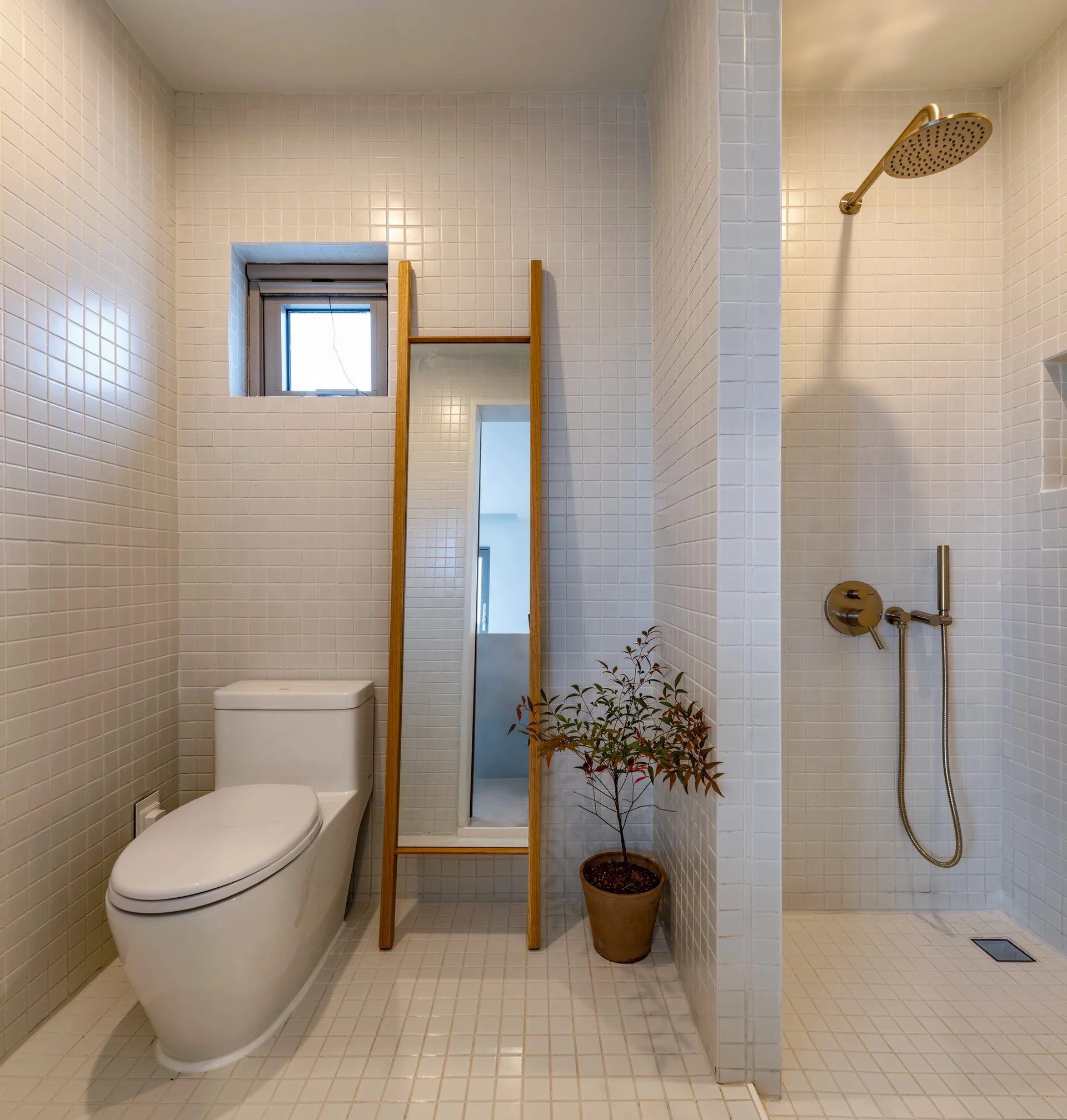 A minimalist bathroom with white square tiles on the walls and floor, featuring a white toilet, wooden leaning mirror, and brass shower fixtures.