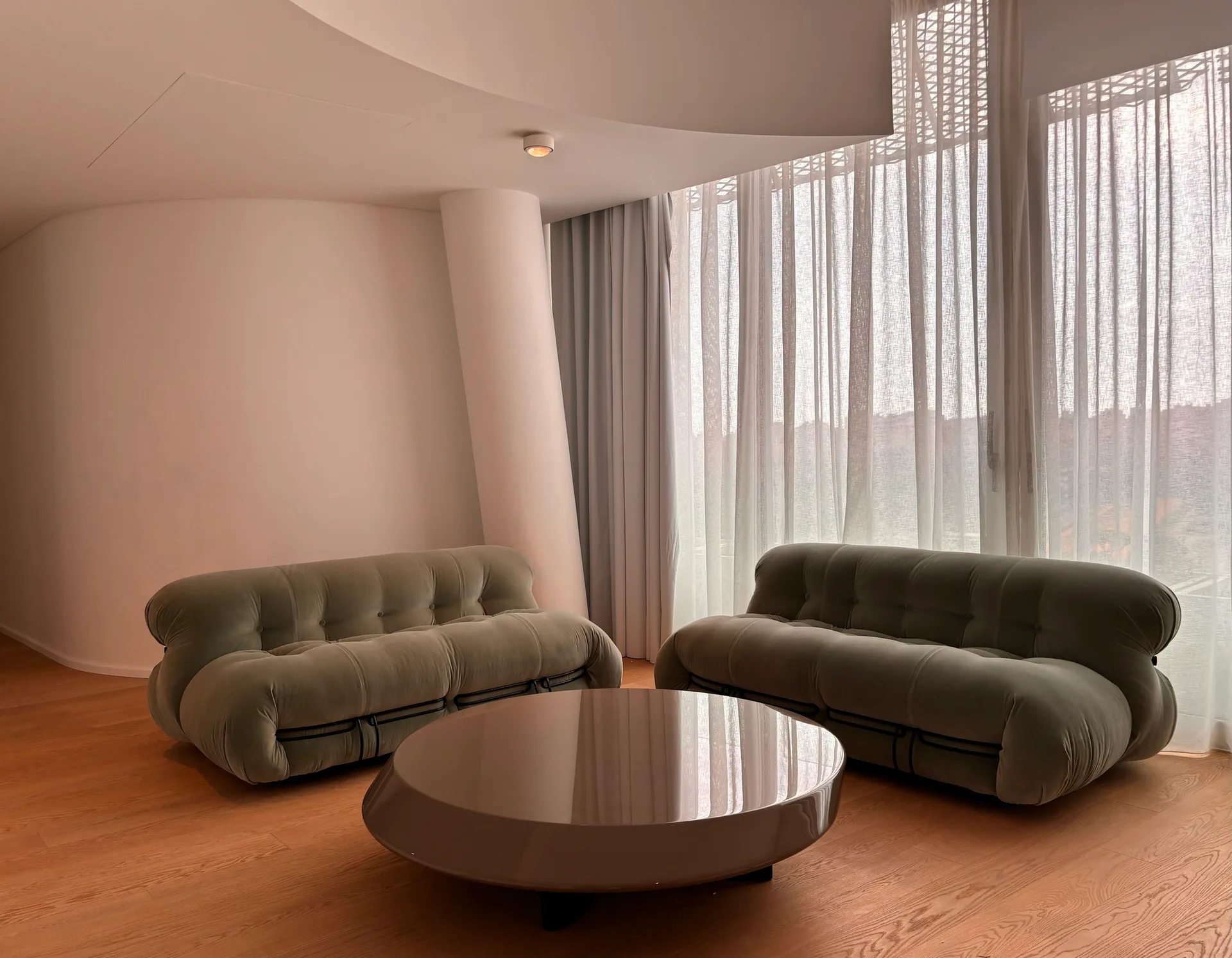 Two olive green tufted lounge sofas sit on a light wood floor near a round glossy coffee table and tall sheer white curtains.