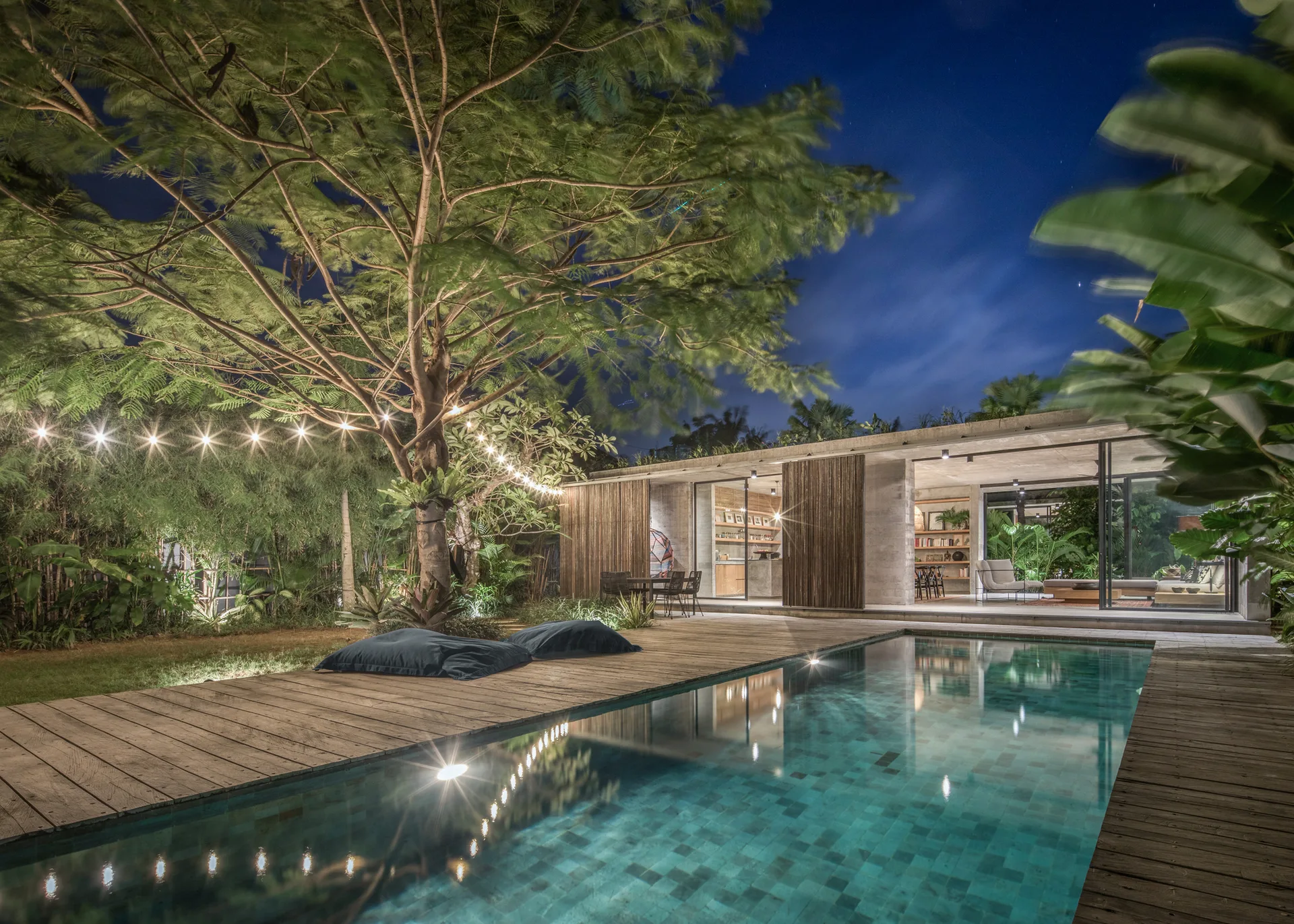 A rectangular swimming pool sits next to a wooden deck and a modern concrete house with sliding glass walls under a lit tree at night.