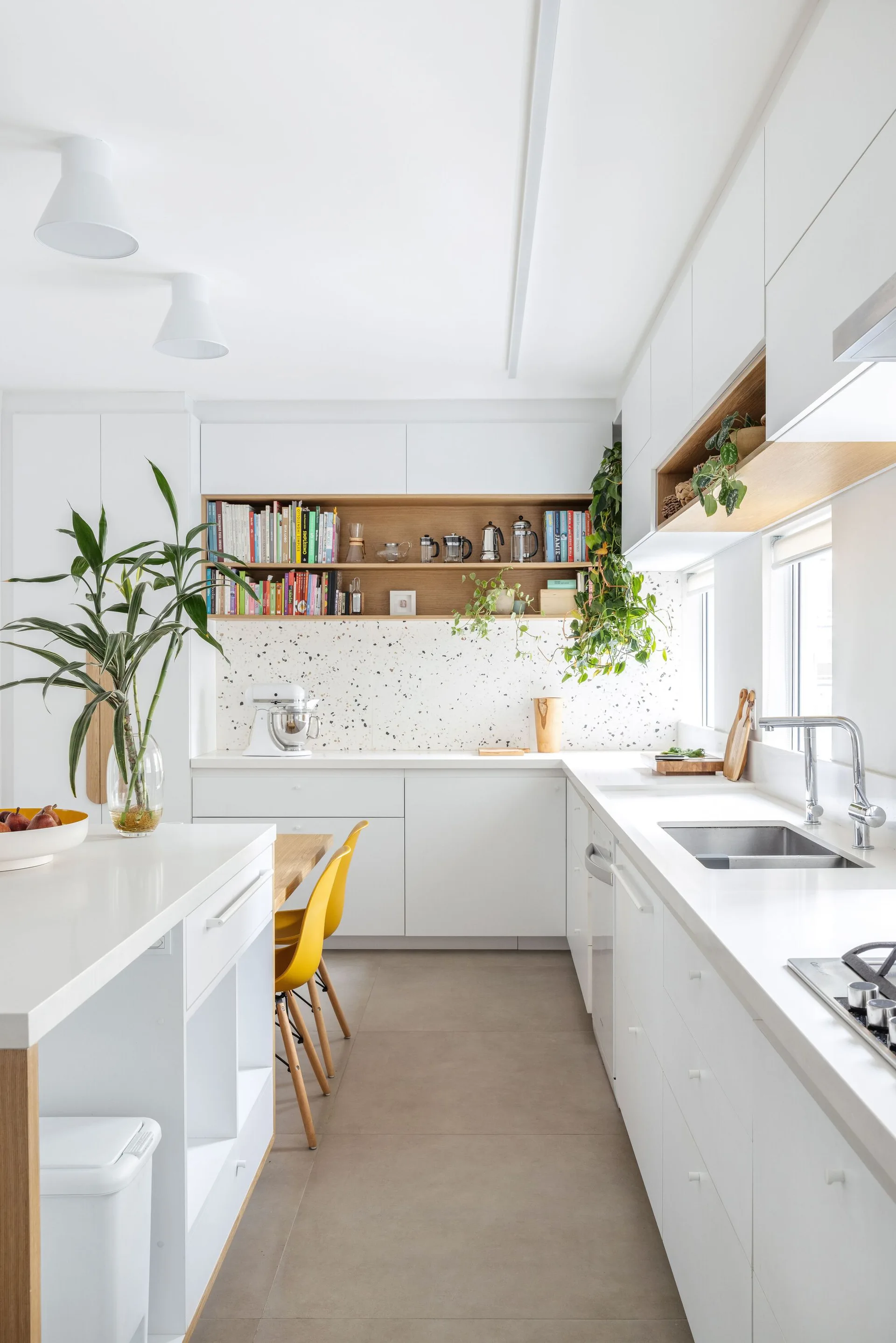 A bright white modern kitchen featuring flat-front cabinets, a speckled terrazzo backsplash, built-in wooden shelving, and yellow dining chairs.