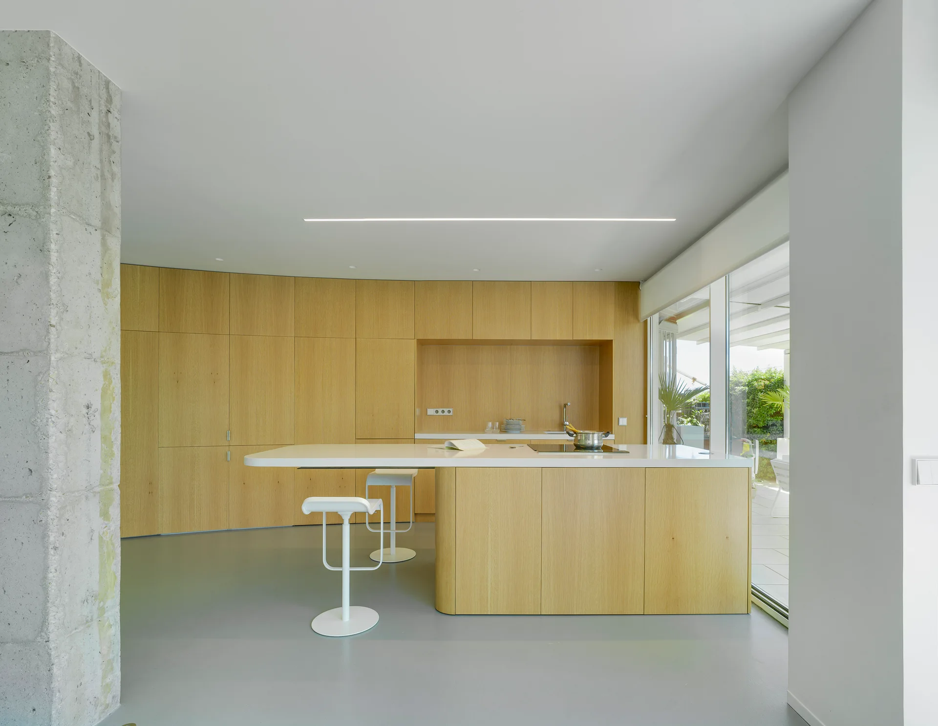 A minimalist kitchen featuring floor-to-ceiling light wood cabinetry, a white countertop island with bar stools, and a raw concrete column.