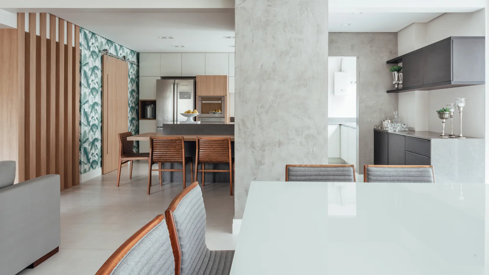 A modern open-plan dining and kitchen area featuring a textured concrete column, a glossy white table, grey chairs, and a wood slat wall.