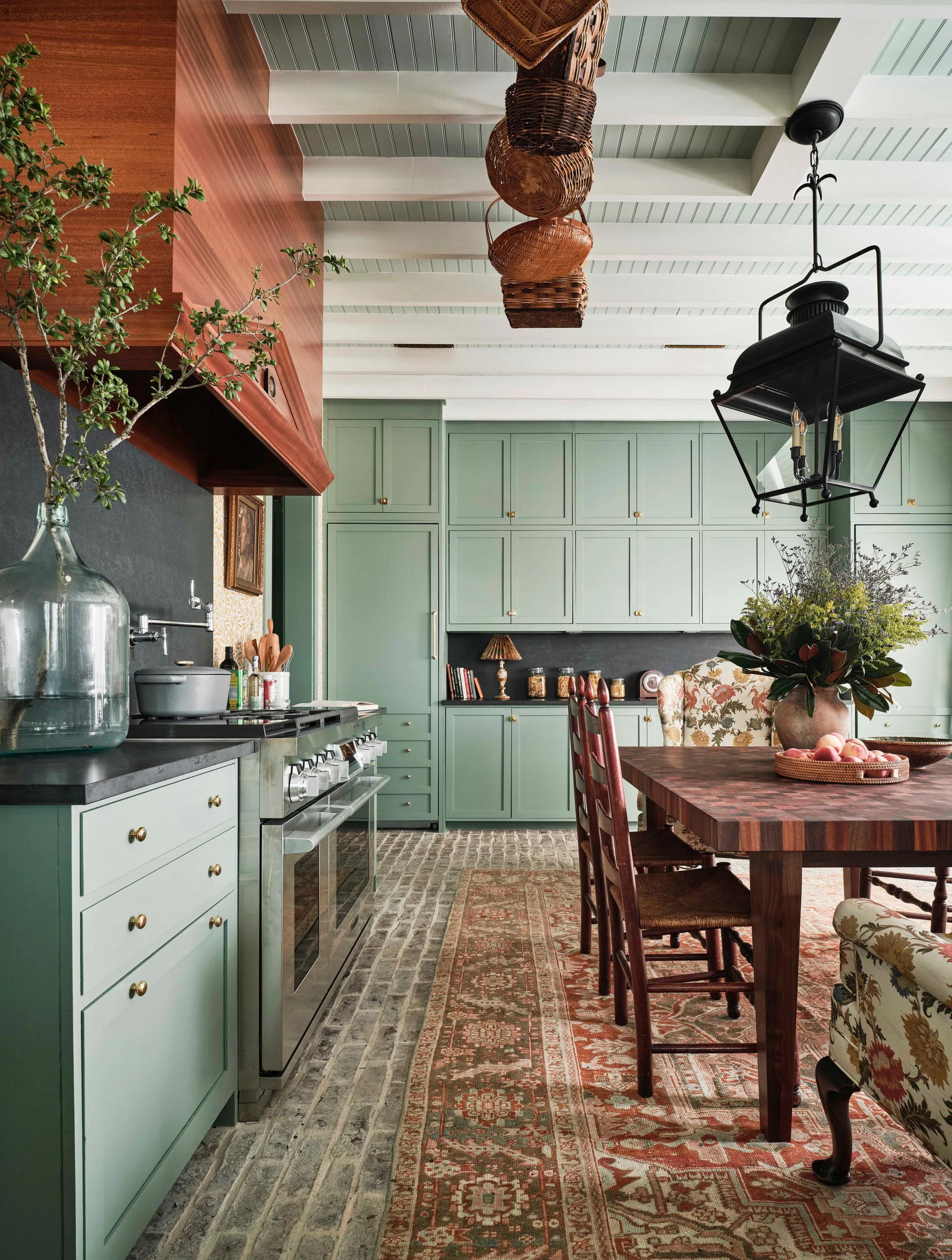 A farmhouse kitchen features sage green cabinetry, a brick floor, a wooden dining table under a black lantern, and a white beamed ceiling.