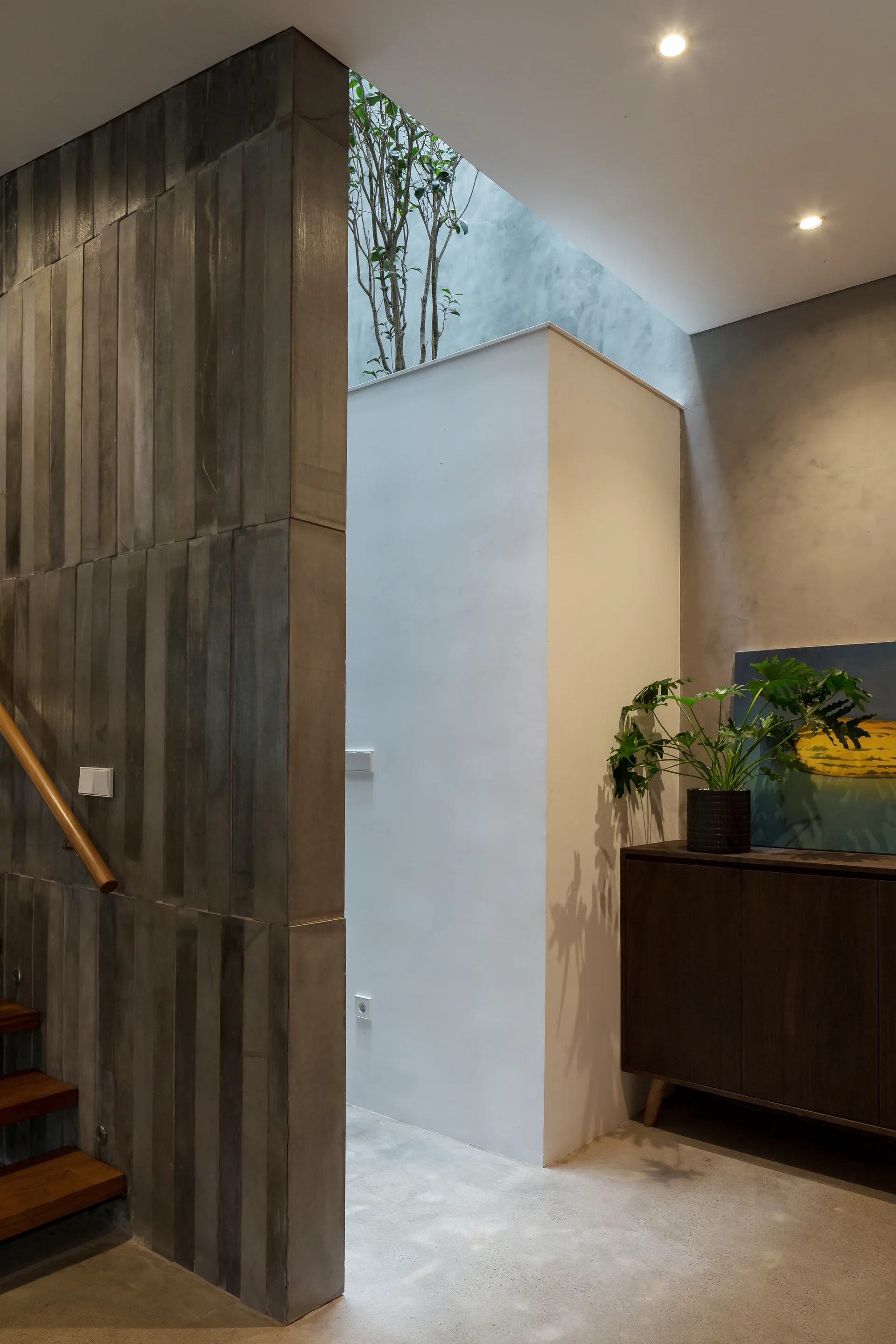 A modern interior space with a textured gray concrete wall, wooden steps, a dark wood console cabinet, and an overhead skylight showing green plants.